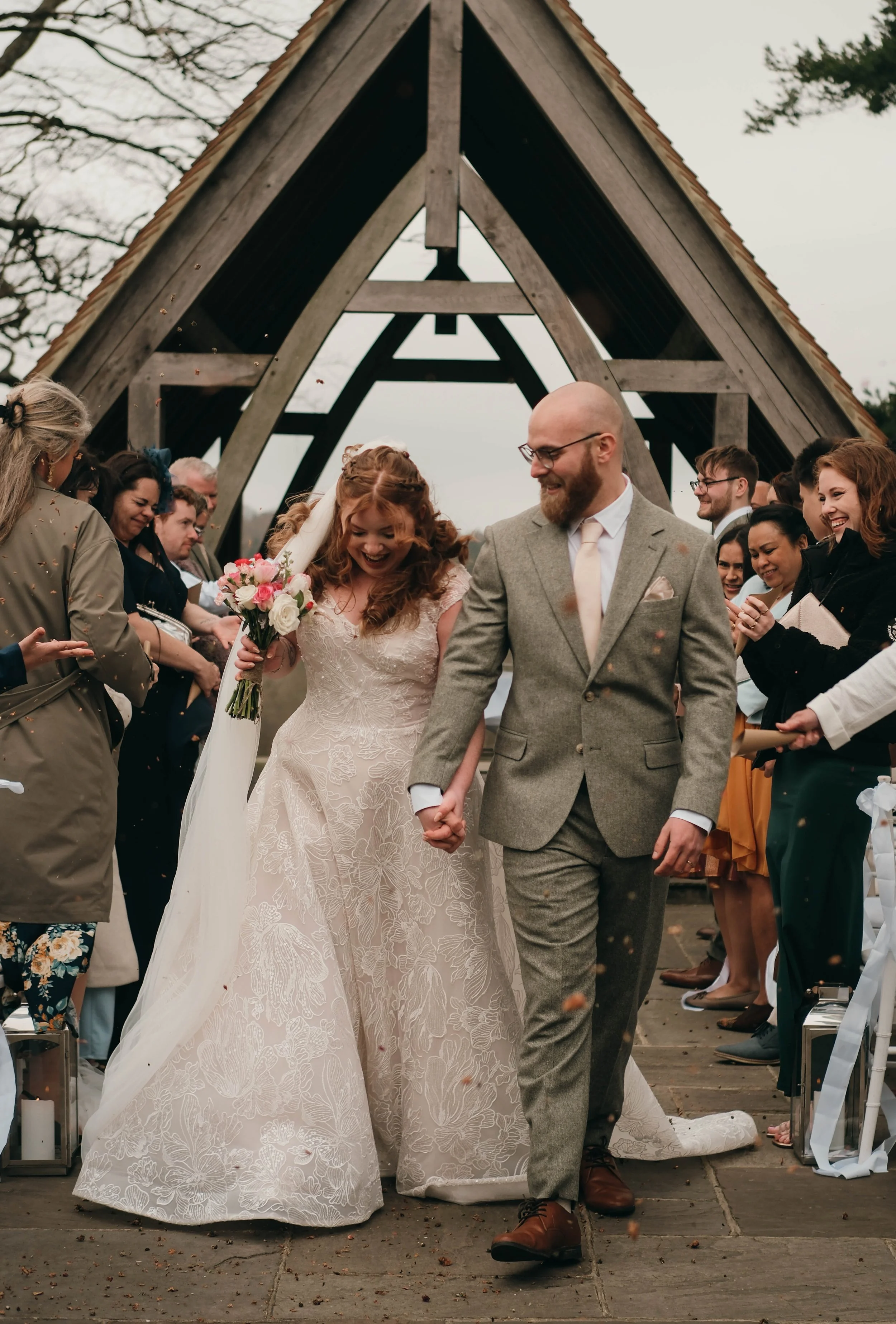 A newlywed couple holding hands and smiling at each other during an outdoor wedding ceremony, with friends and family celebrating around them beneath a wooden A-frame structure.