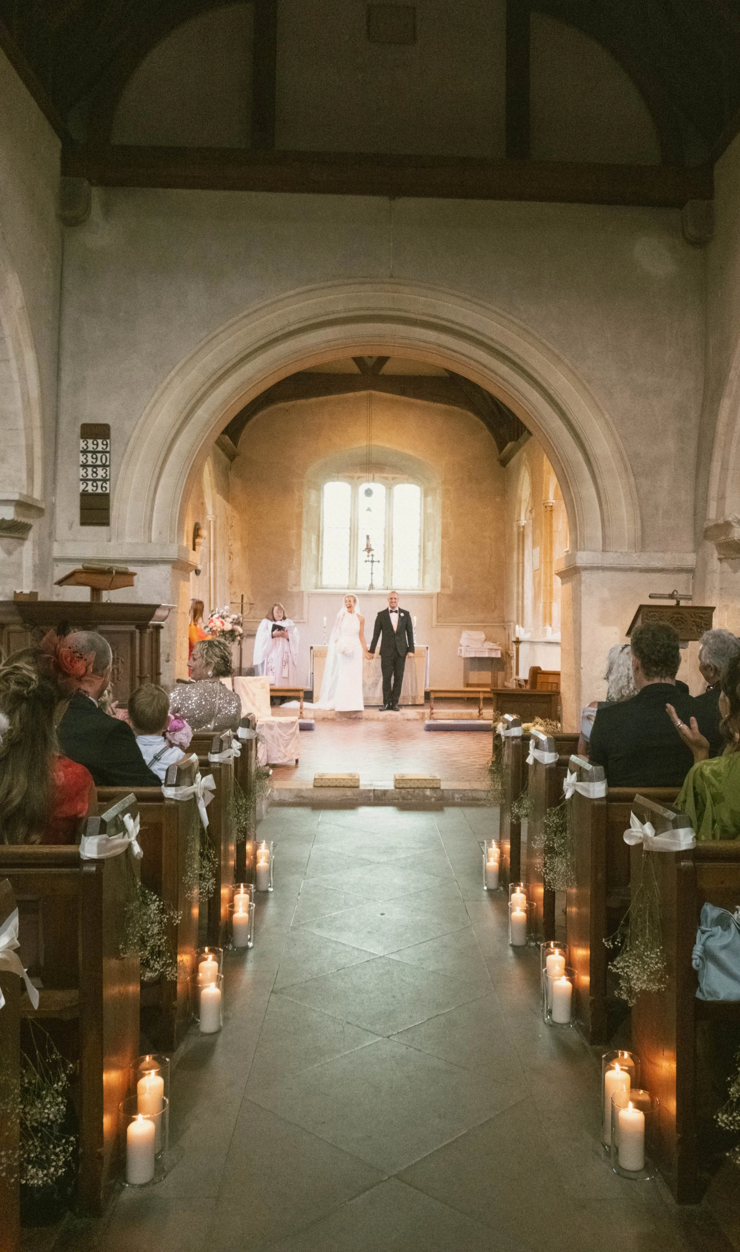 A wedding ceremony inside a church with the bride and groom holding hands in front of an altar. Guests are seated in pews decorated with white ribbons and flowers, and candles line the aisle.