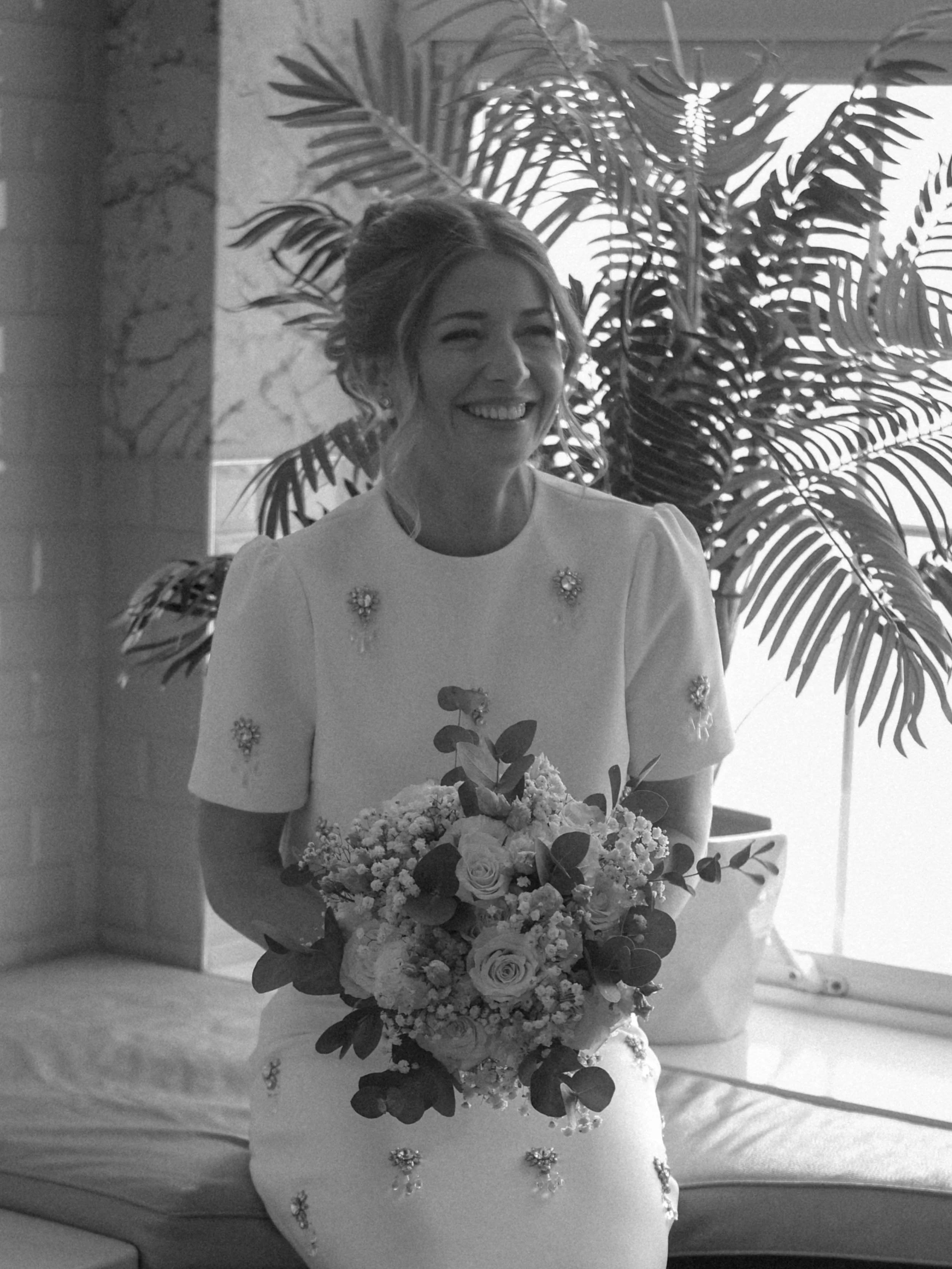 A woman smiling and holding a bouquet of flowers, standing in front of a large leafy plant and a marble wall.
