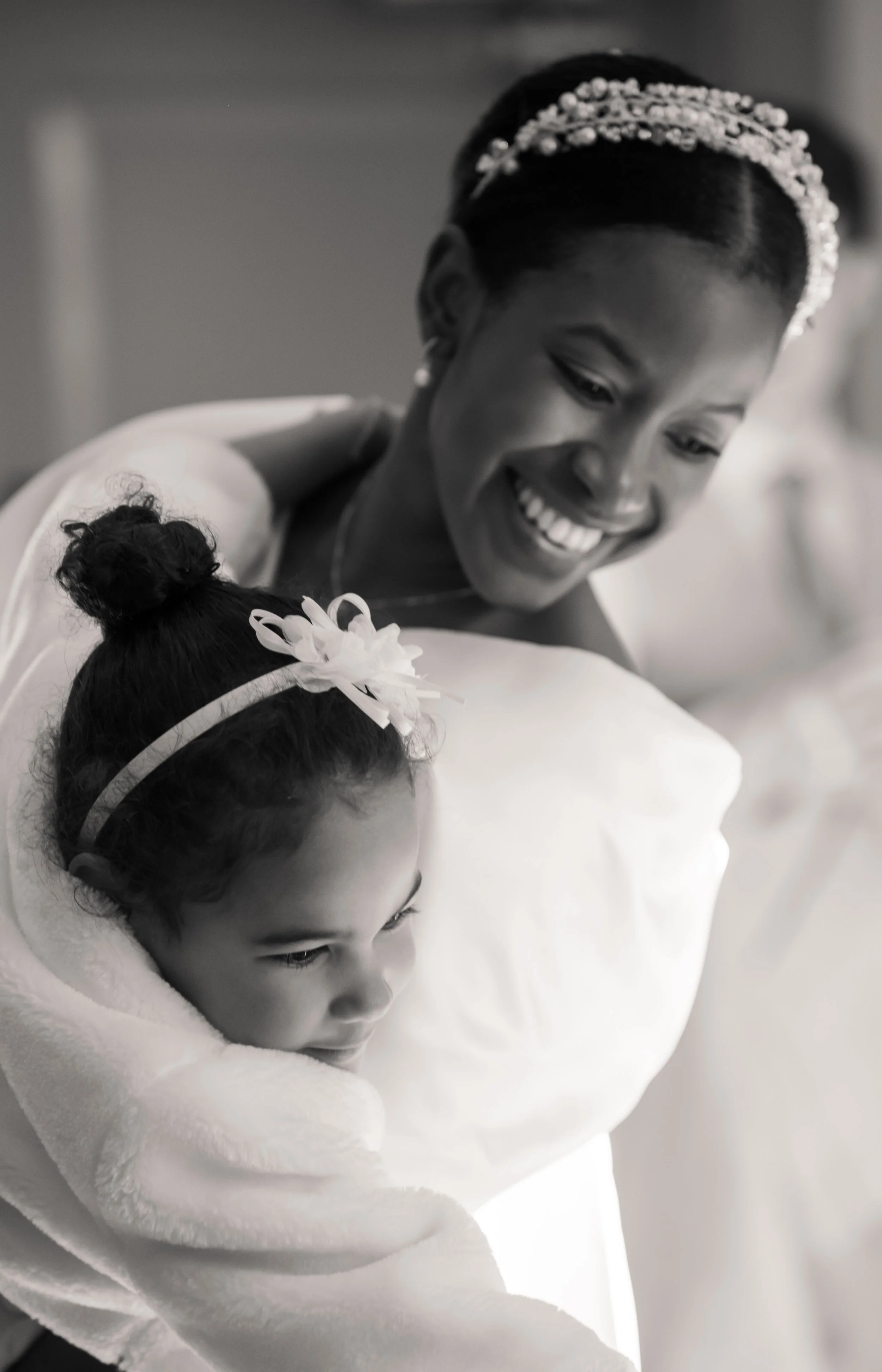 A woman with an elegant hairstyle wearing a tiara and smiling, embracing a child in a white robe with a flower hair accessory.