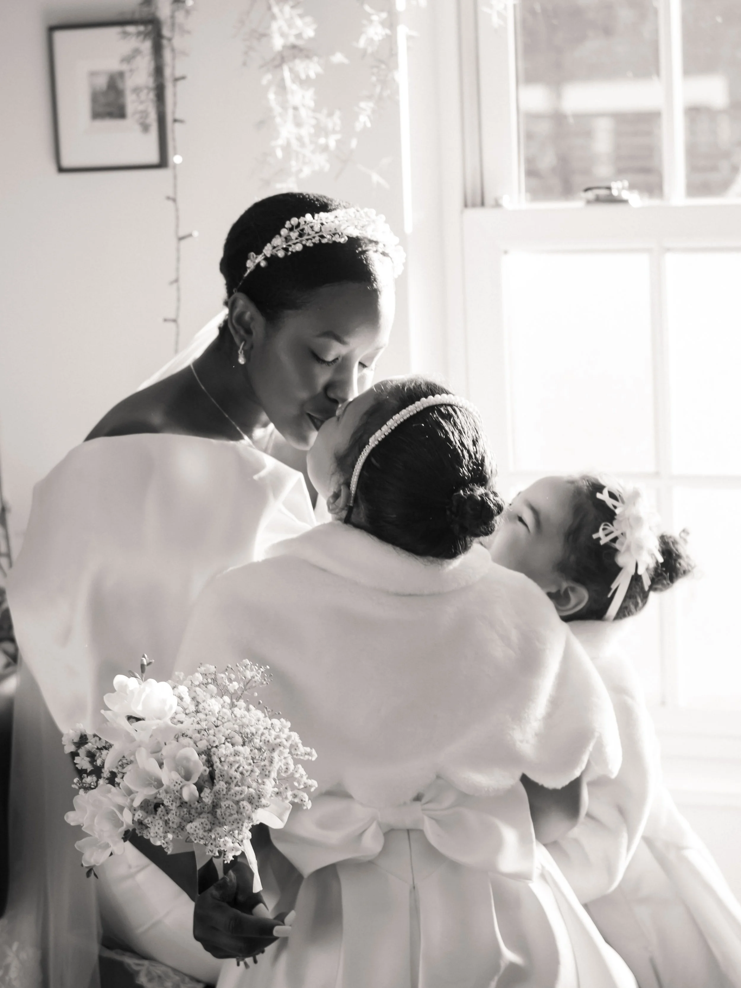 A black and white photo of two women, dressed in wedding gowns, sharing a kiss. One woman is holding a bouquet of flowers. The scene is illuminated by sunlight coming through a window, with a framed picture and fairy lights on the wall in the backgro