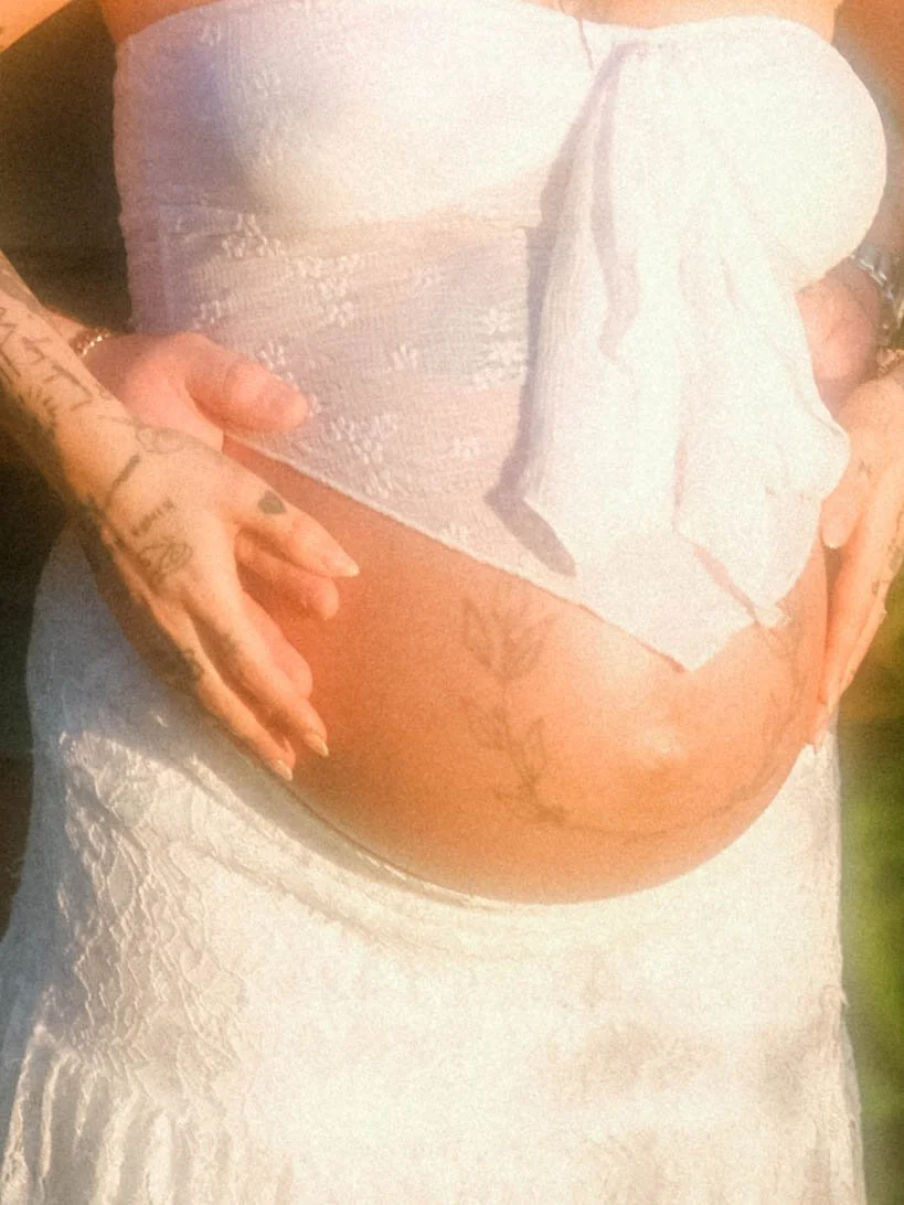A close-up of a pregnant woman wearing a white dress and holding her belly with both hands, showing tattoos on her left hand and belly.