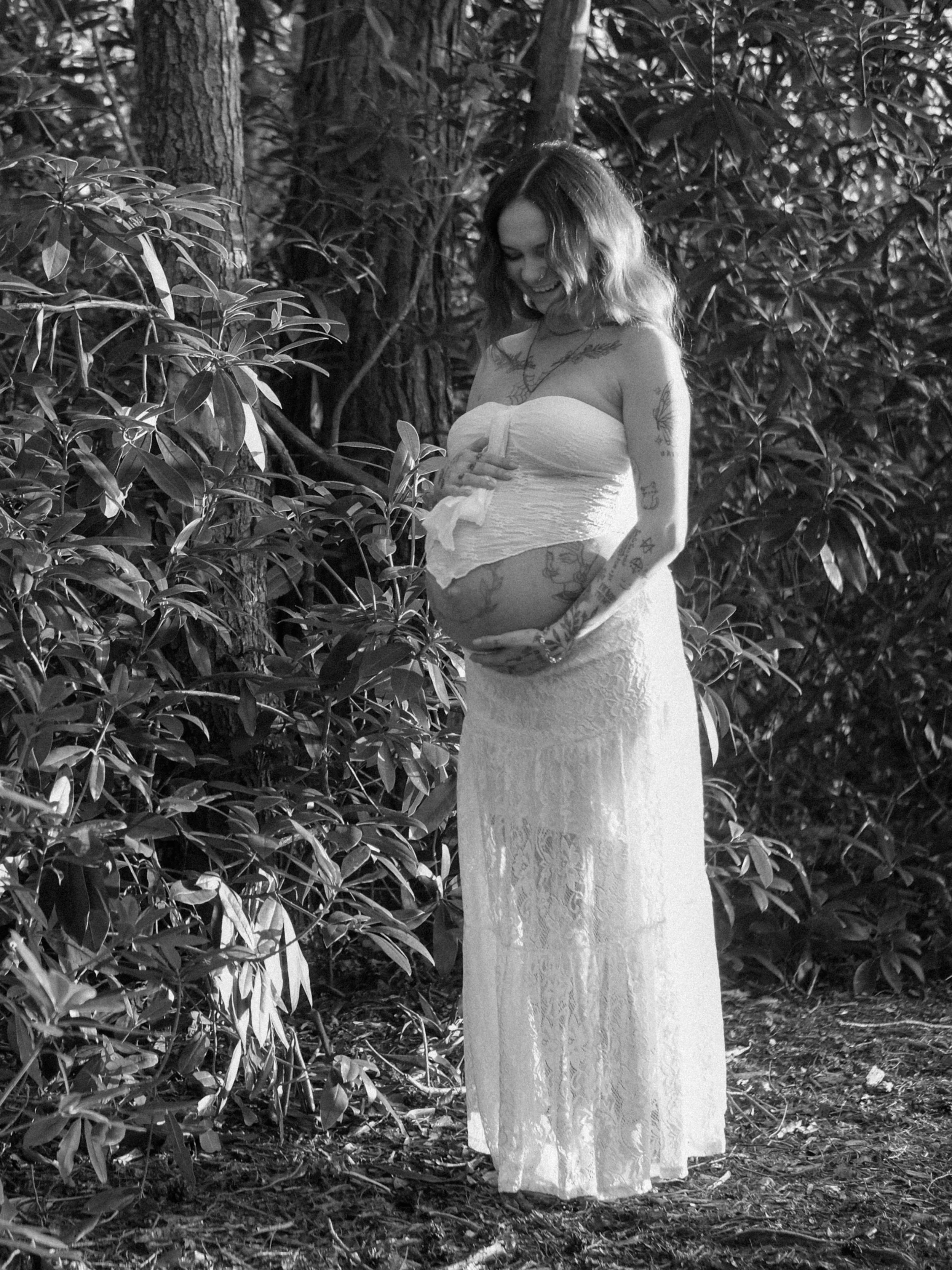 A smiling woman standing outdoors in a forested area, with one hand resting on her pregnant belly. She is wearing a strapless dress and has tattoos on her arms and chest, with wavy hair and surrounded by trees and foliage.