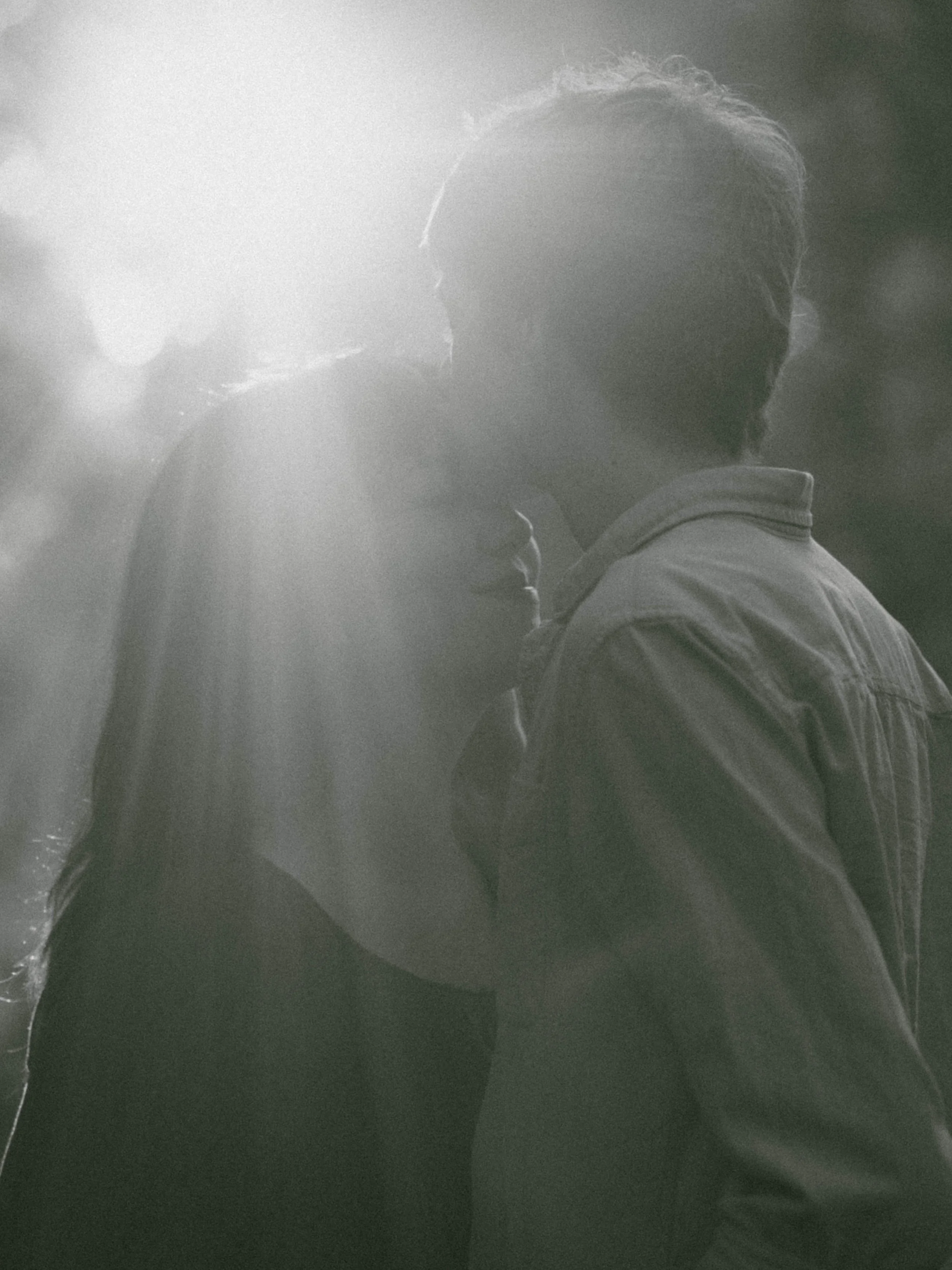 A person in profile with braided hair, wearing a jacket, standing opposite another person in a hooded jacket, sharing a tender moment in black and white.