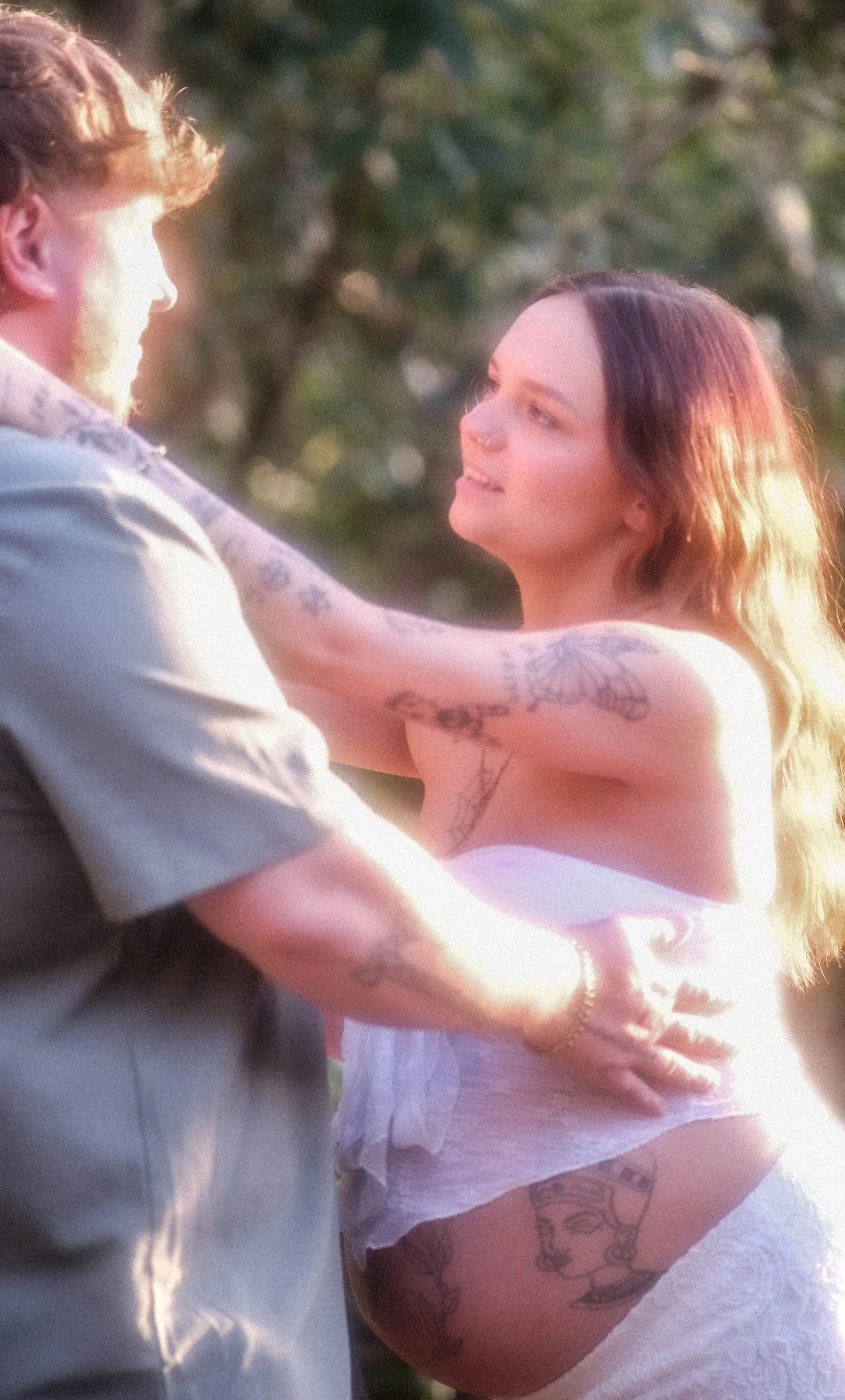 A young woman with dark hair and tattoos on her arm and thigh, wearing a white dress, looking at a man with a beard and tattoos on his arm, outdoors during sunset.