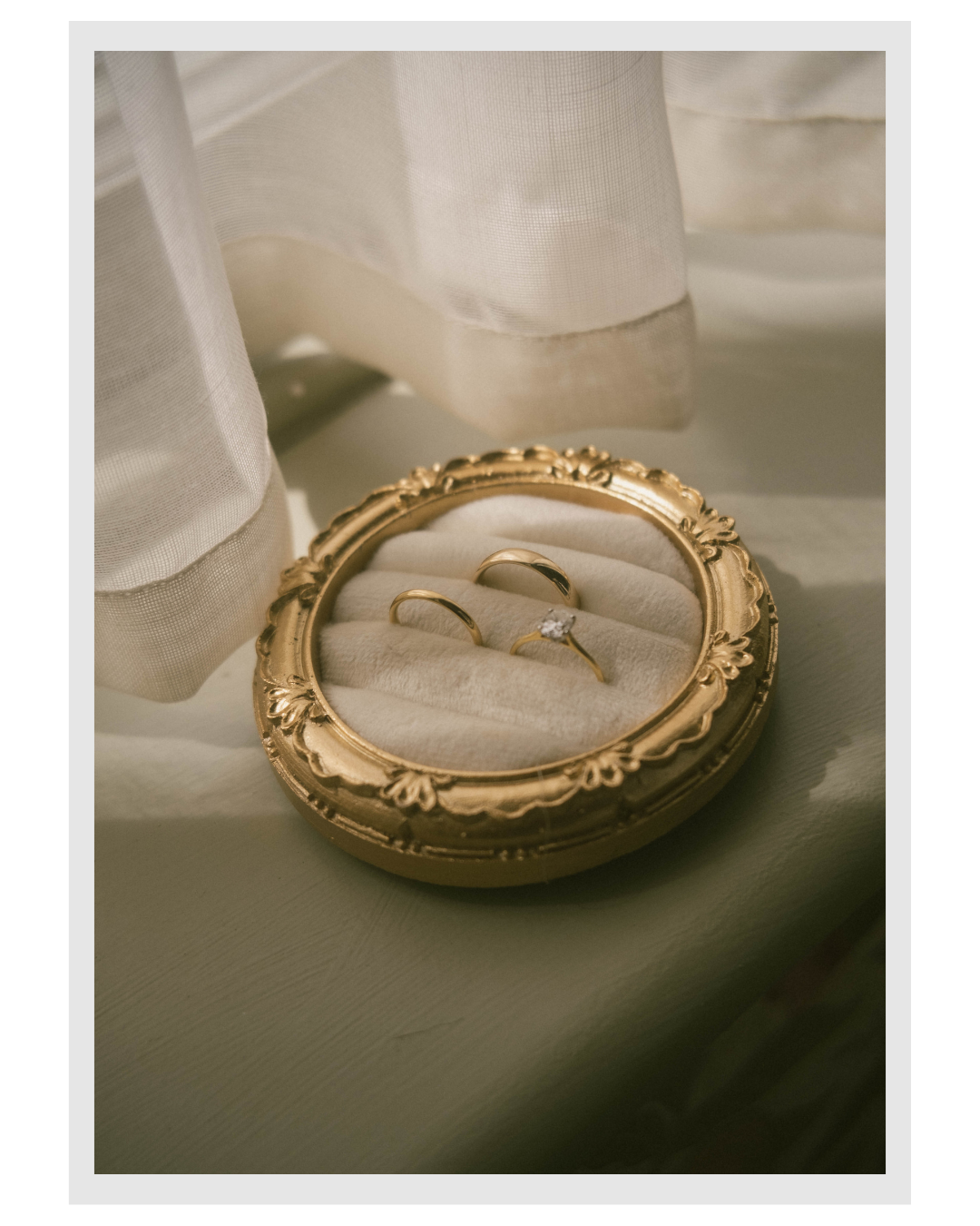 Gold ring box with three gold rings, including one with a diamond, on a white velvet cushion, surrounded by sheer white fabric.
