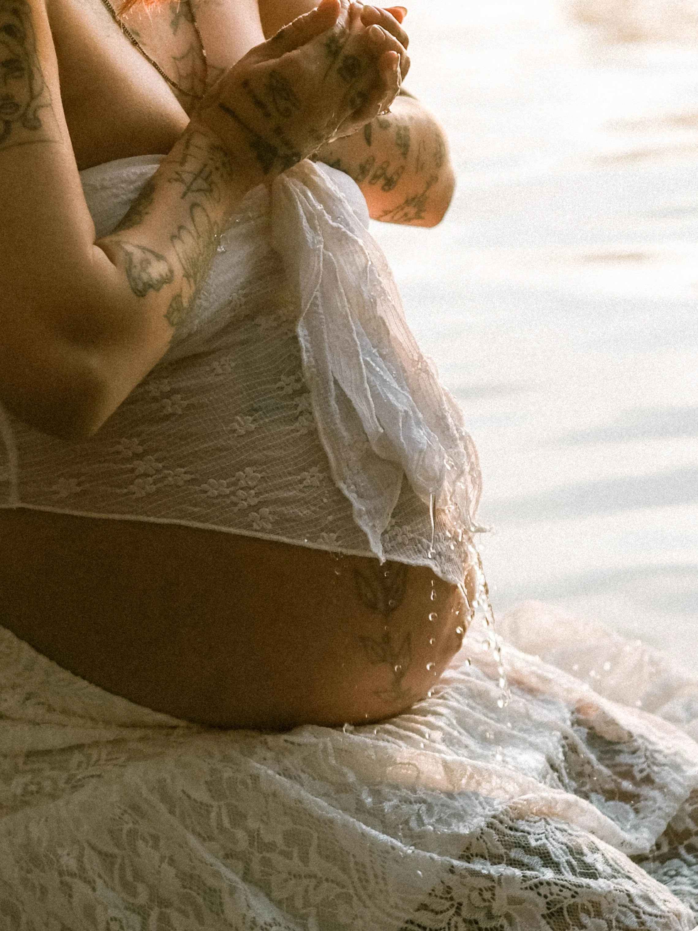 Close-up of a person sitting on a lace-covered surface, holding their hands together, with tattoos visible on their arms, wearing a white lace garment. The background is a light-colored, softly lit environment.