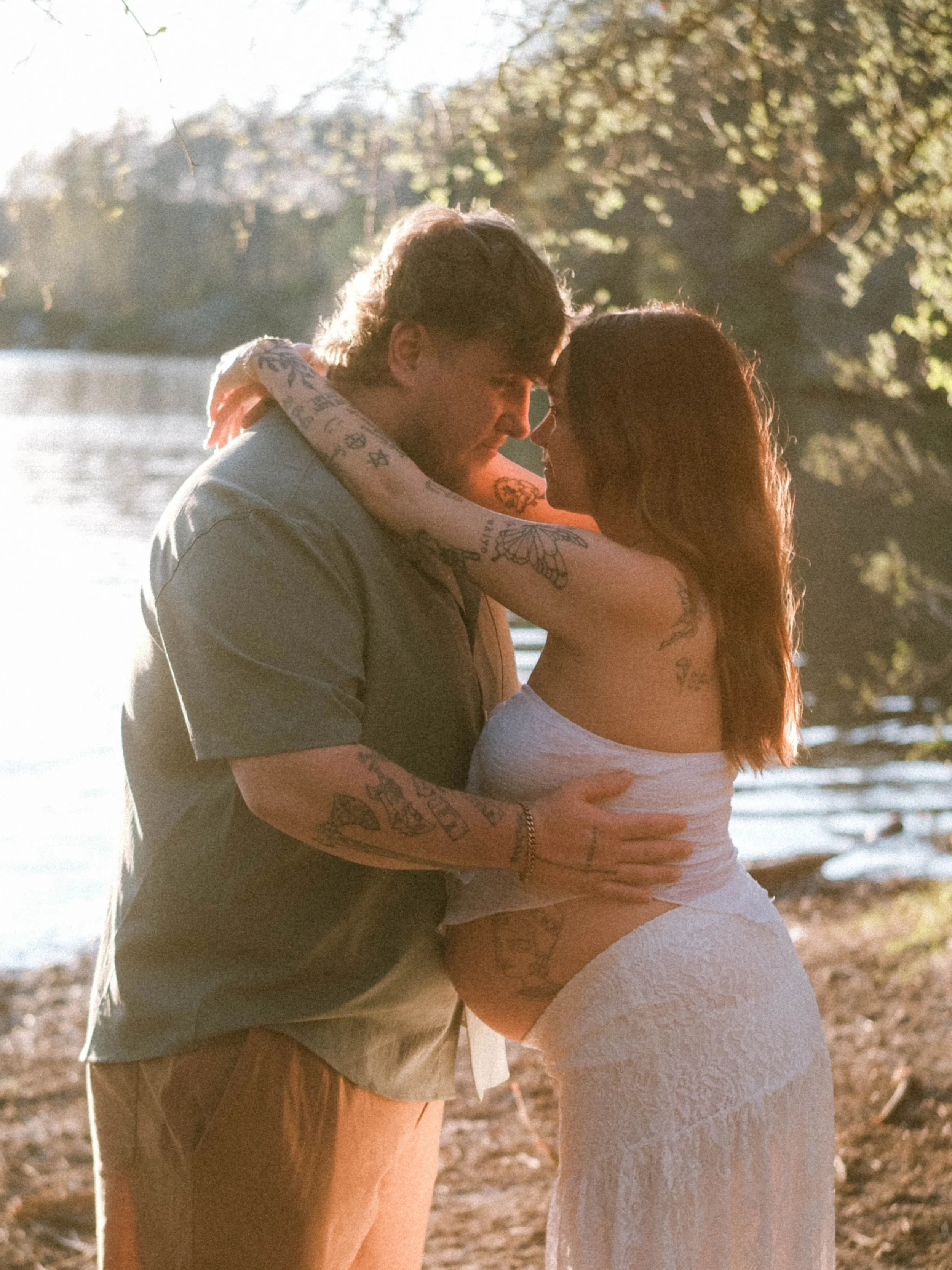 A couple embraces on a beach during sunset, with trees and a body of water in the background.