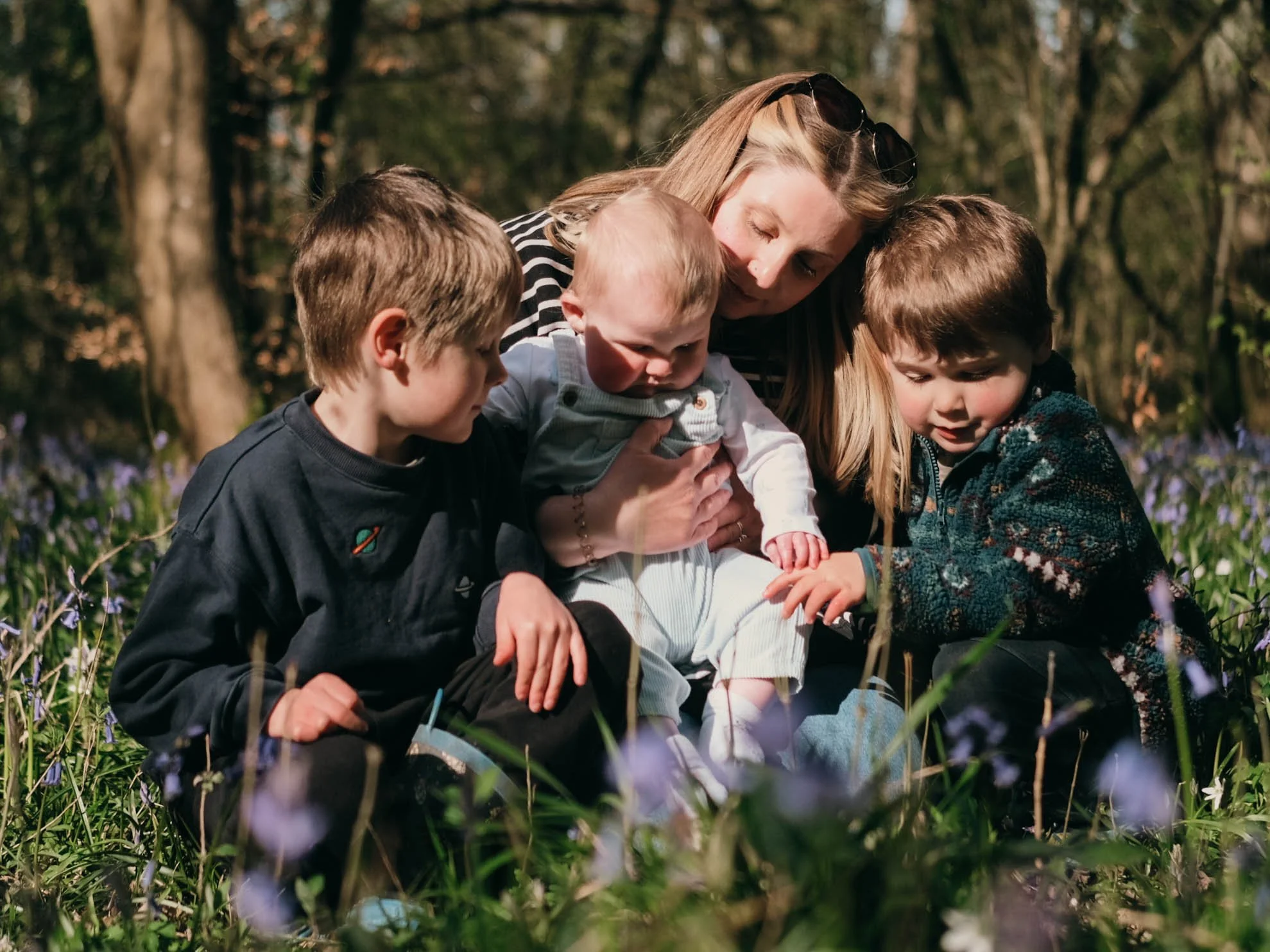 A woman and four young children sitting outdoors in a wooded area surrounded by purple flowers, engaging in a close, affectionate moment.