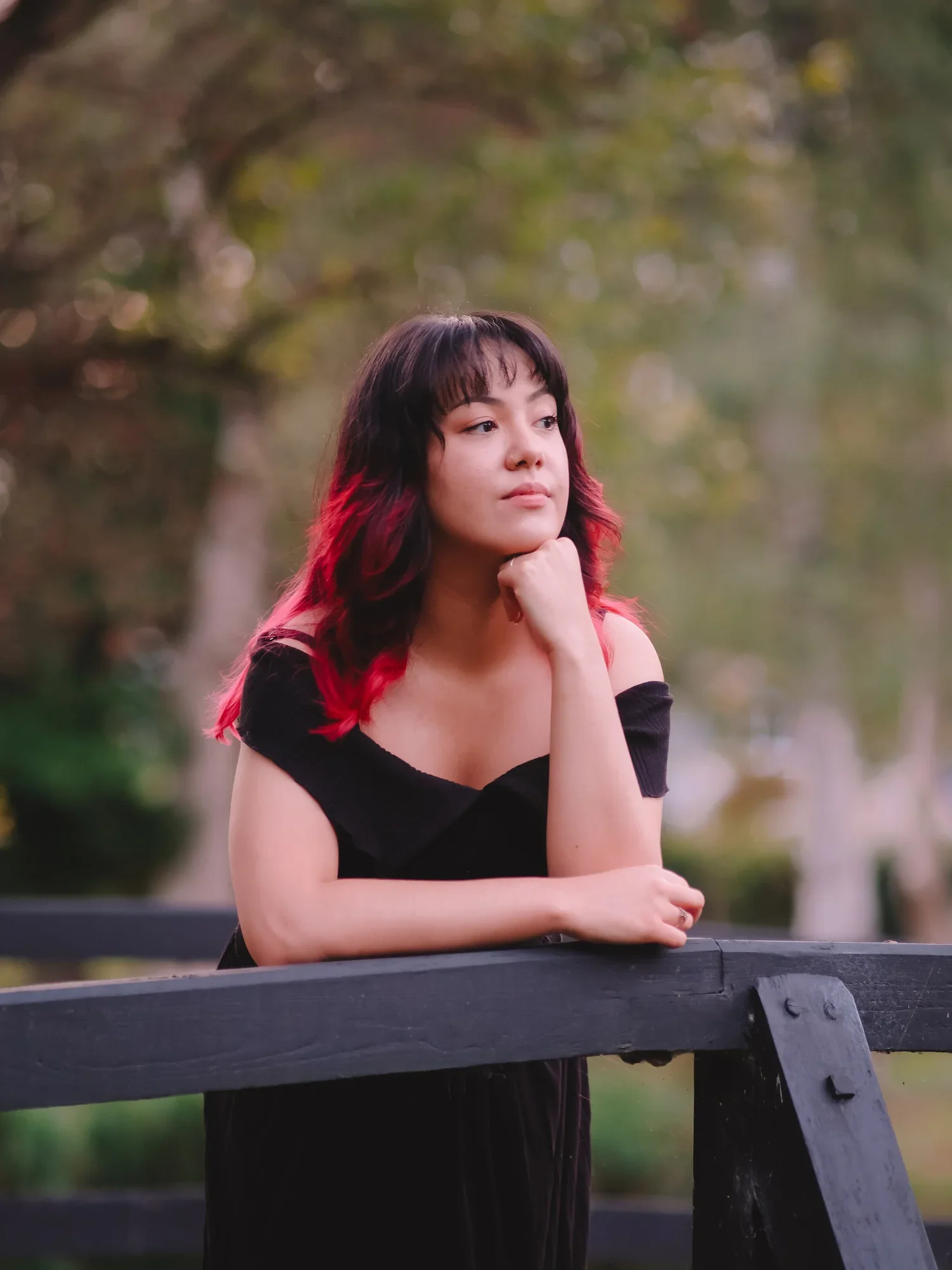 A young woman with dark hair and red tips, wearing a black off-shoulder top, sitting on a park bench with her chin resting on her hand, looking thoughtfully into the distance, with a blurred green and yellow background of trees.