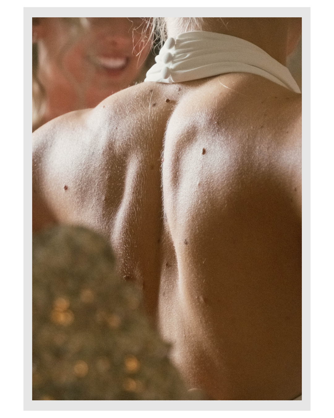 Close-up of a woman's bare back and shoulder, with a smiling face partially visible, wearing a white top or dress with a bow detailing near the shoulder.