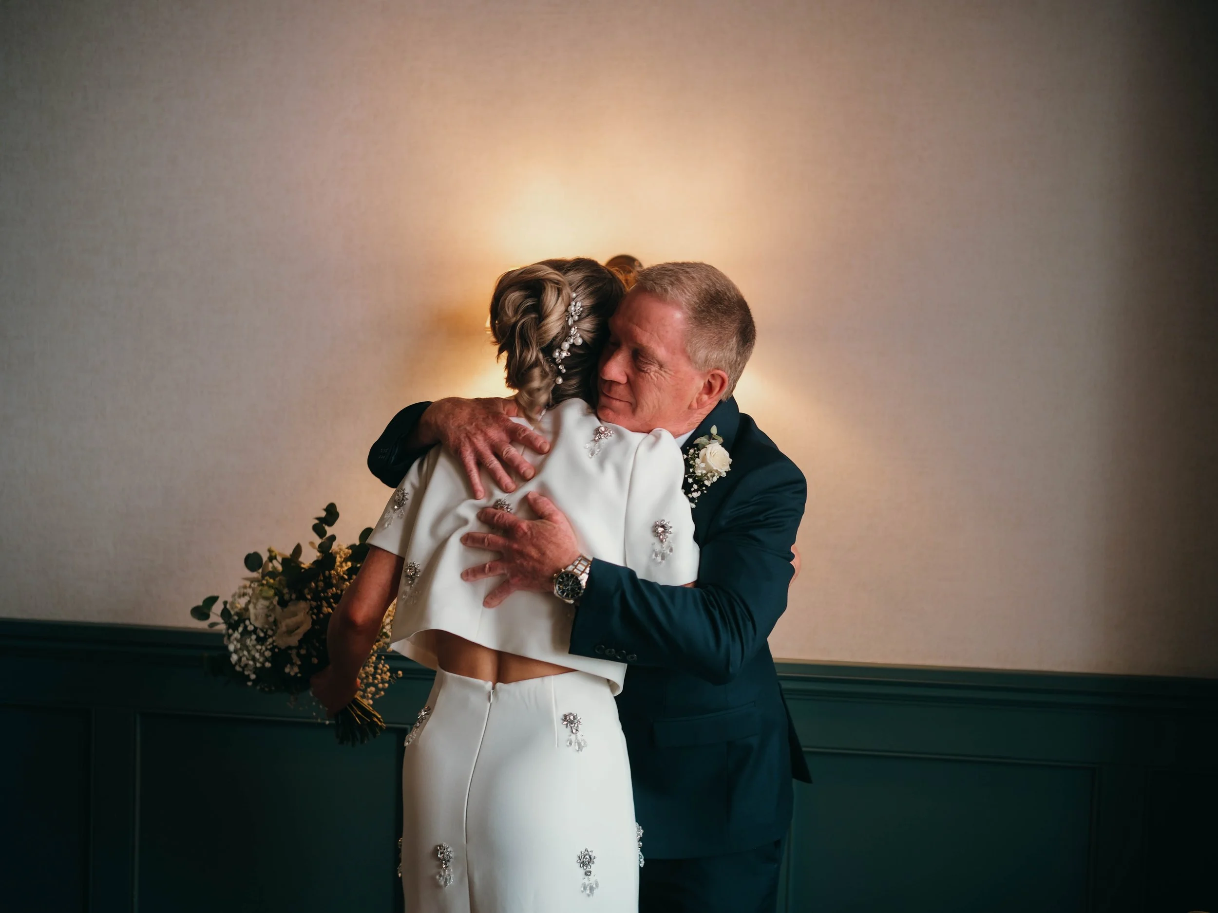 An older man and a woman are sharing a warm hug indoors, with soft lighting and a neutral-colored wall in the background.
