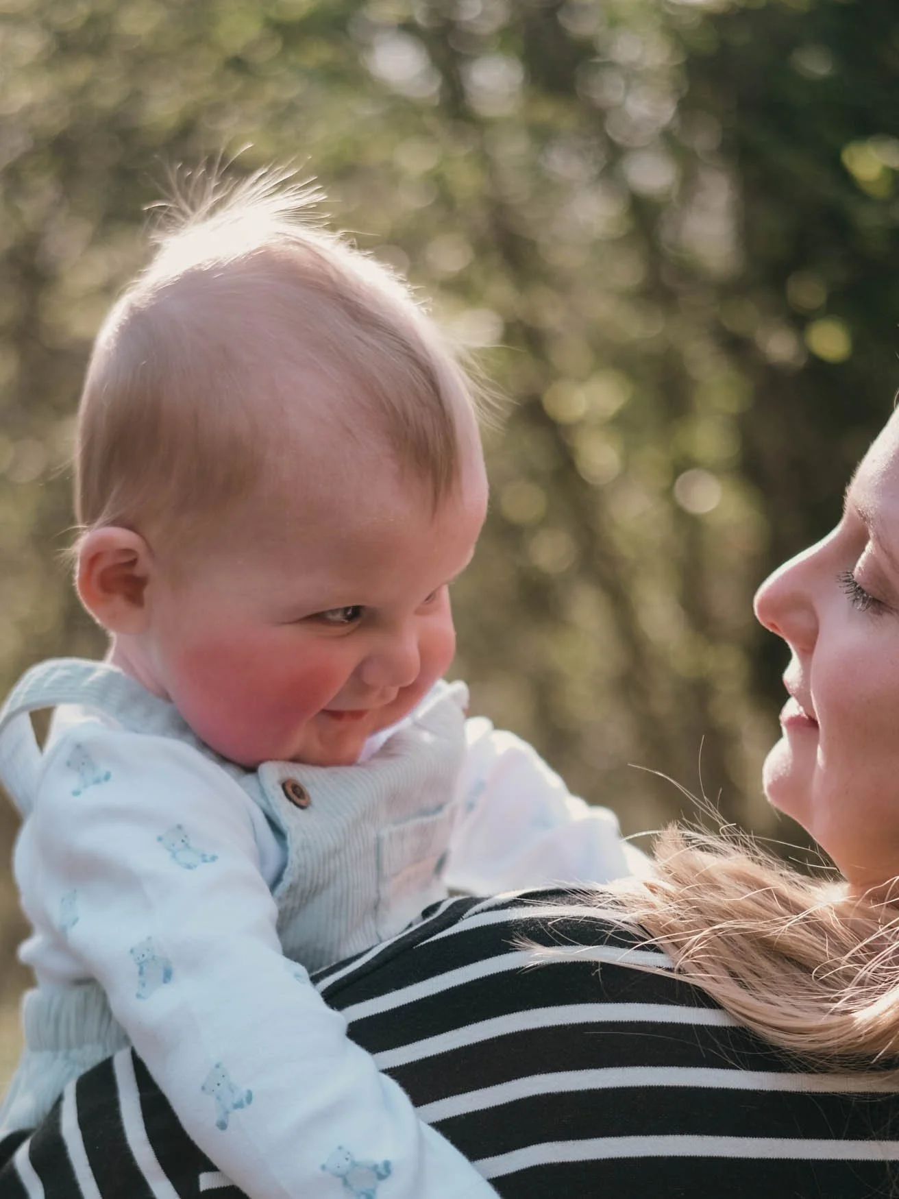 A woman holding a smiling baby outside in sunlight, with trees in the background.