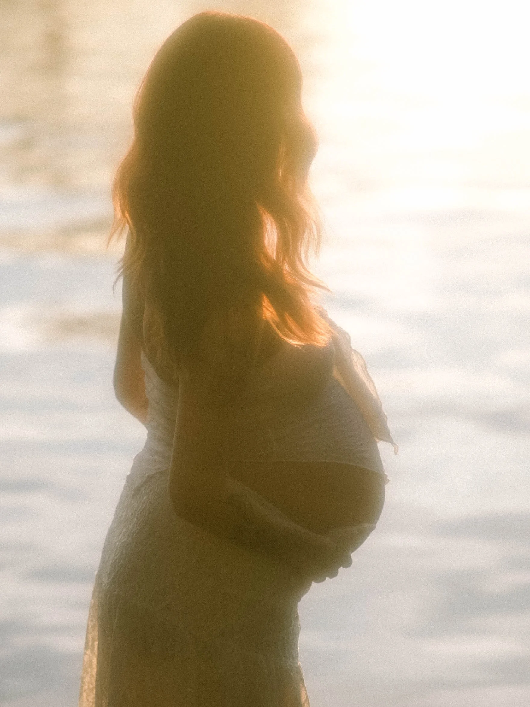 Silhouette of a woman sitting by the water at sunset or sunrise, with long wavy hair and wearing a dress.