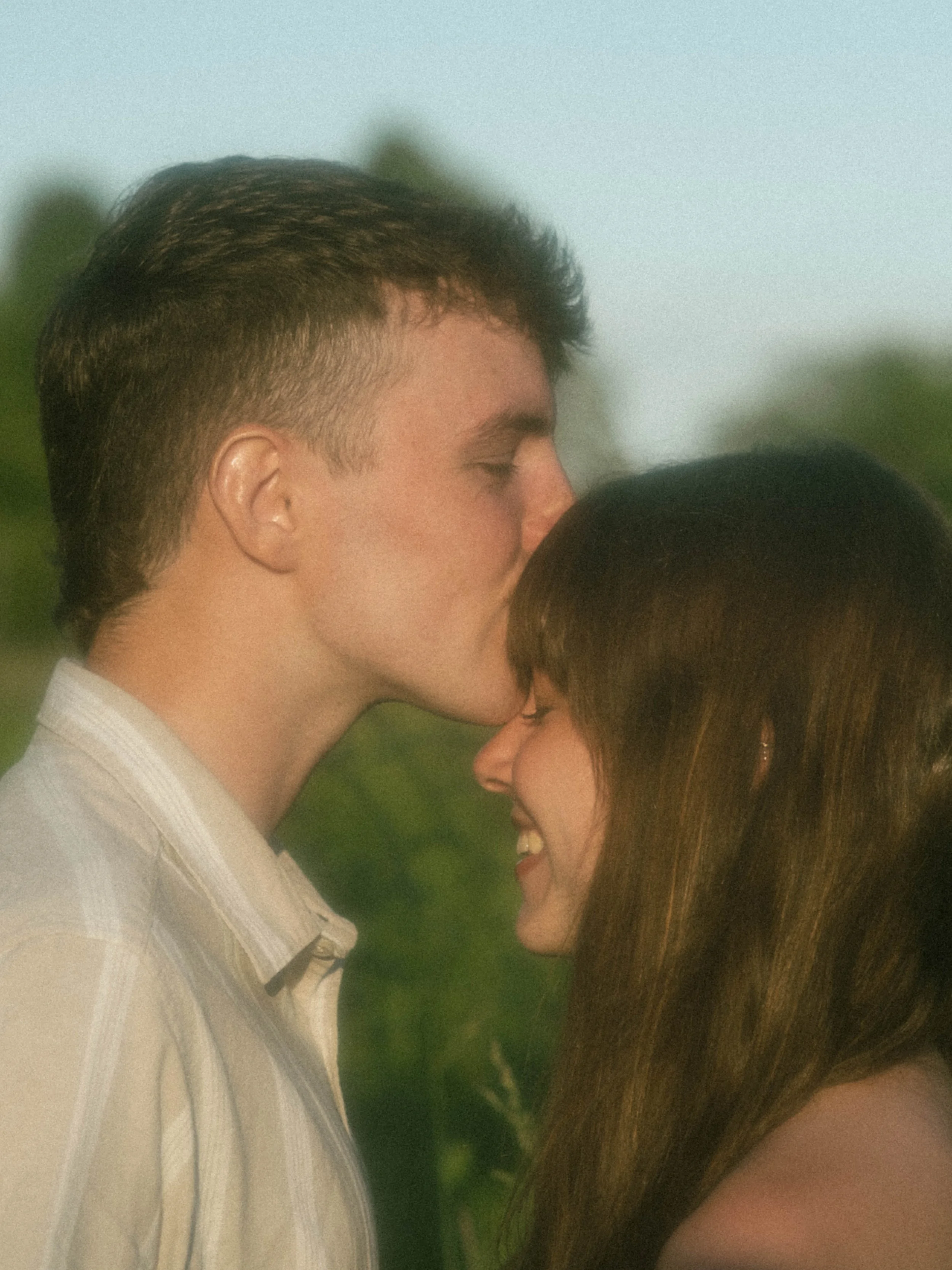 A young man and woman close together, sharing a tender moment with their foreheads touching and eyes closed outdoors with a blurry green background.