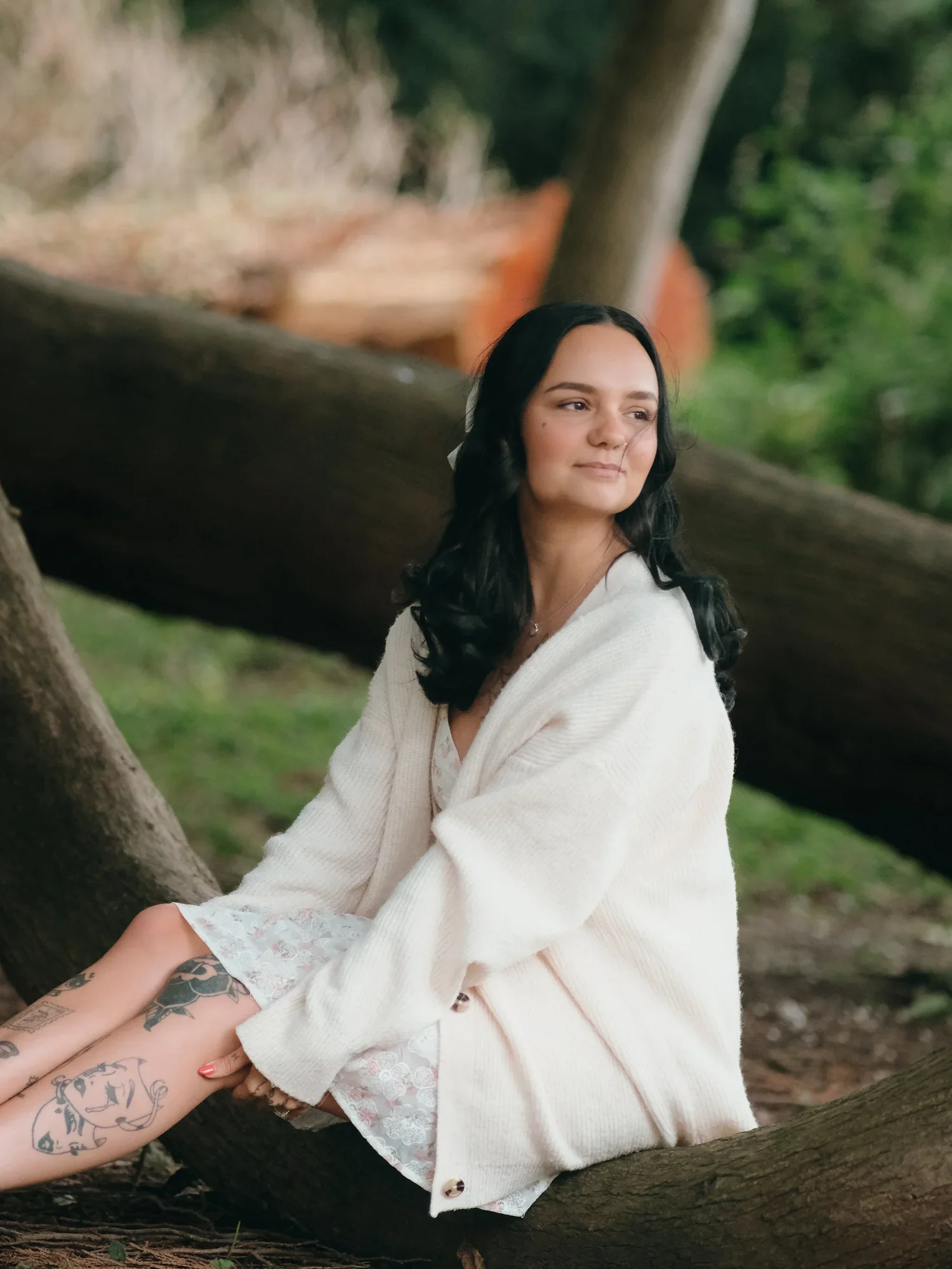 A woman with black hair, wearing a white cardigan, sitting on a tree branch outdoors, with greenery in the background.