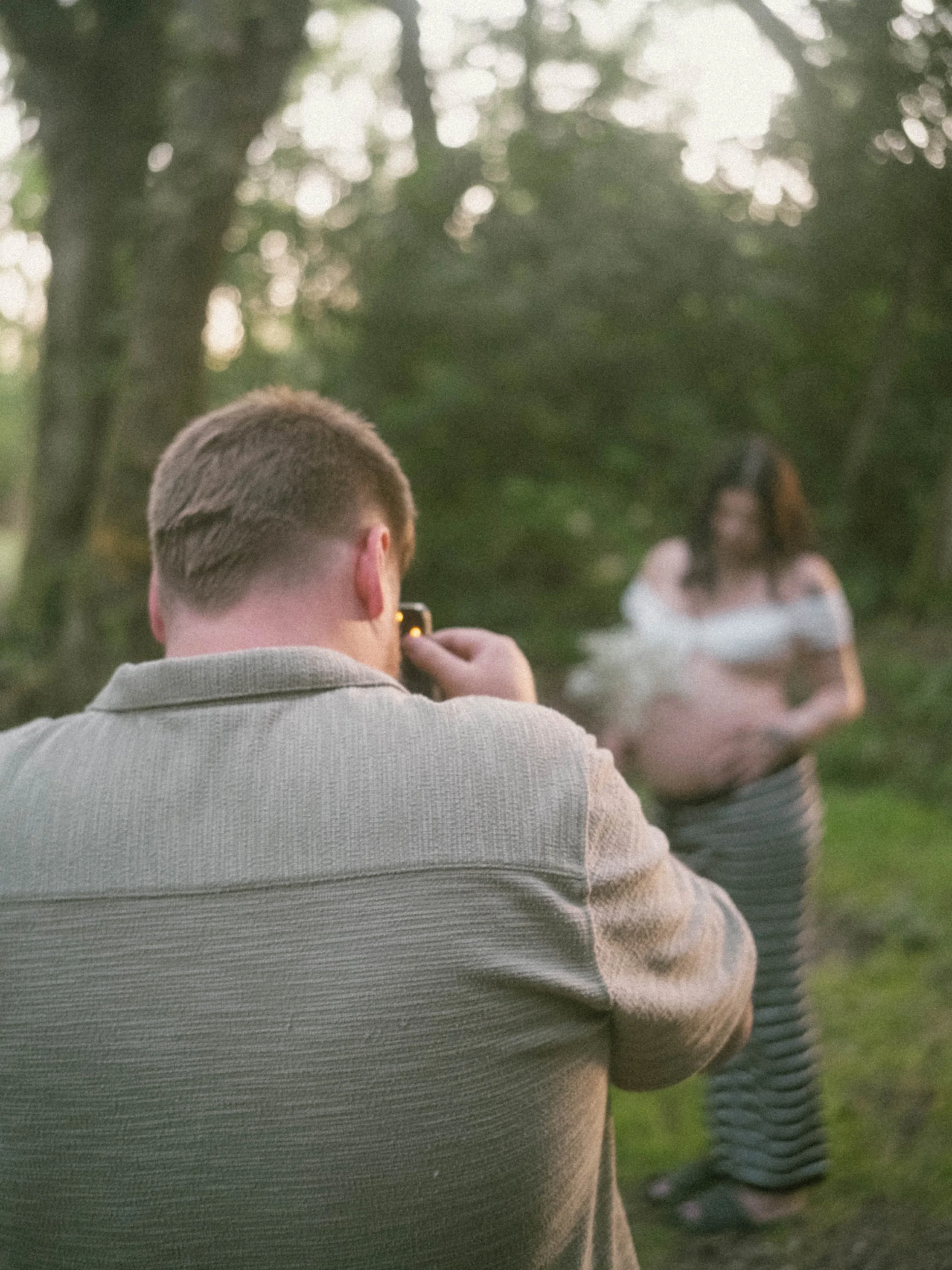 A man with short hair wearing a light-colored sweater, sitting outdoors in a forested area during sunset, capturing a woman in a light dress that is sitting on the ground.