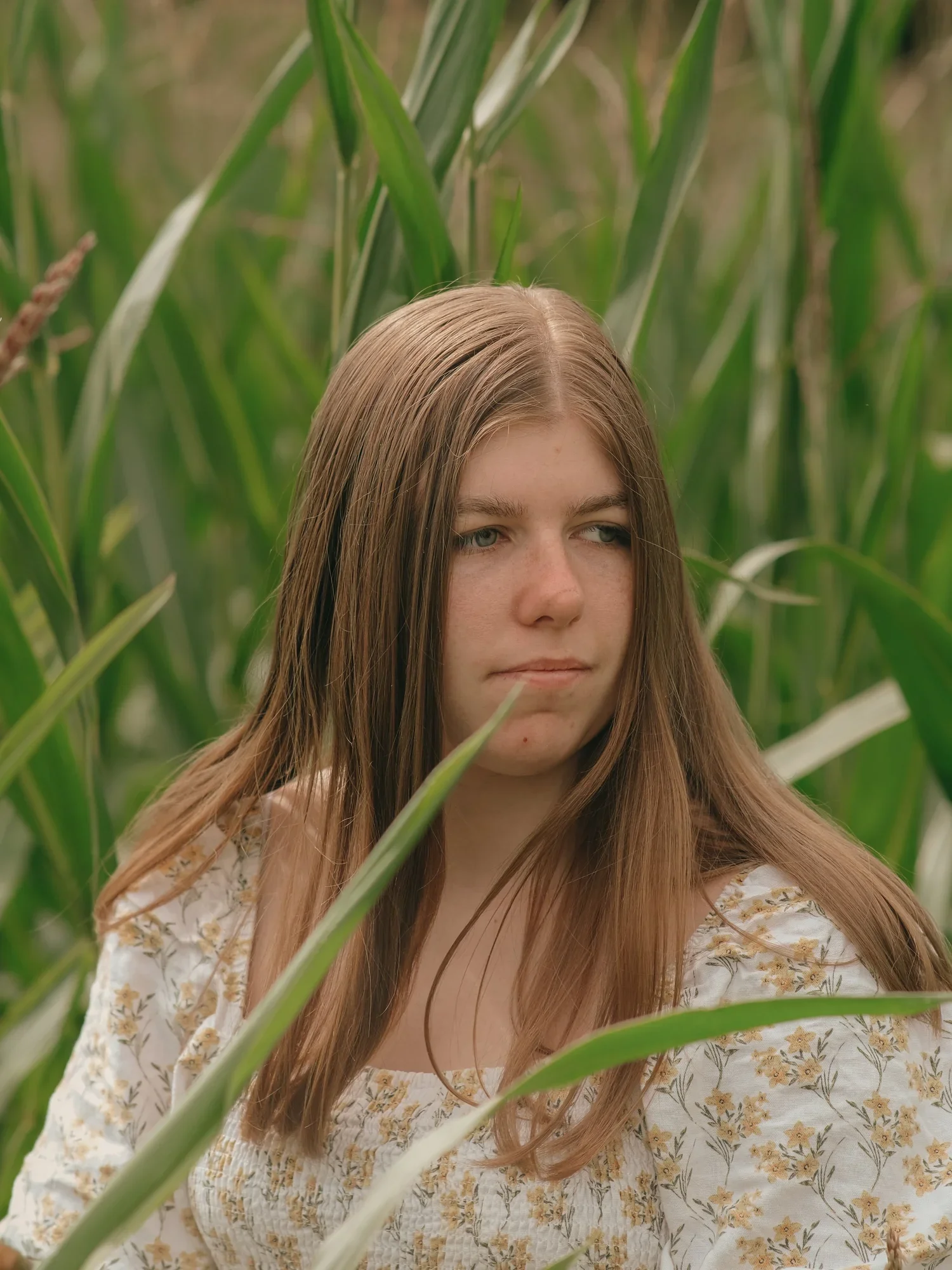 A young woman with long, light brown hair and freckles, wearing a white dress with a floral pattern, standing amidst tall green plants, possibly in a field or garden.