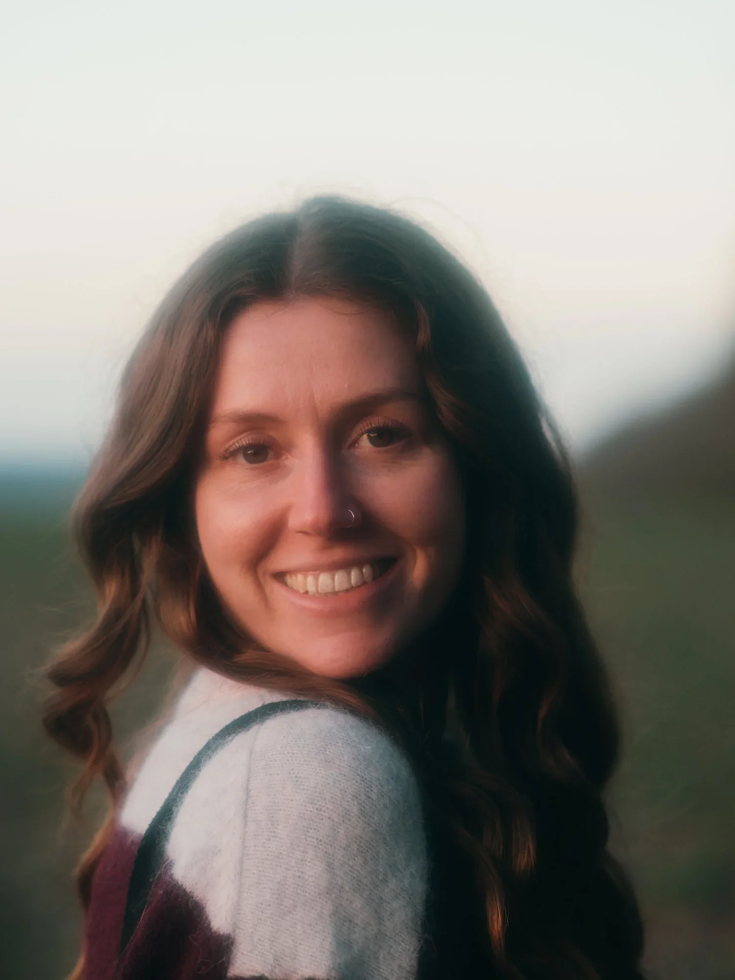 A smiling woman with wavy brown hair and freckles, wearing a white sweater with maroon and green stripes on the sleeve, outdoors with a blurred landscape in the background.