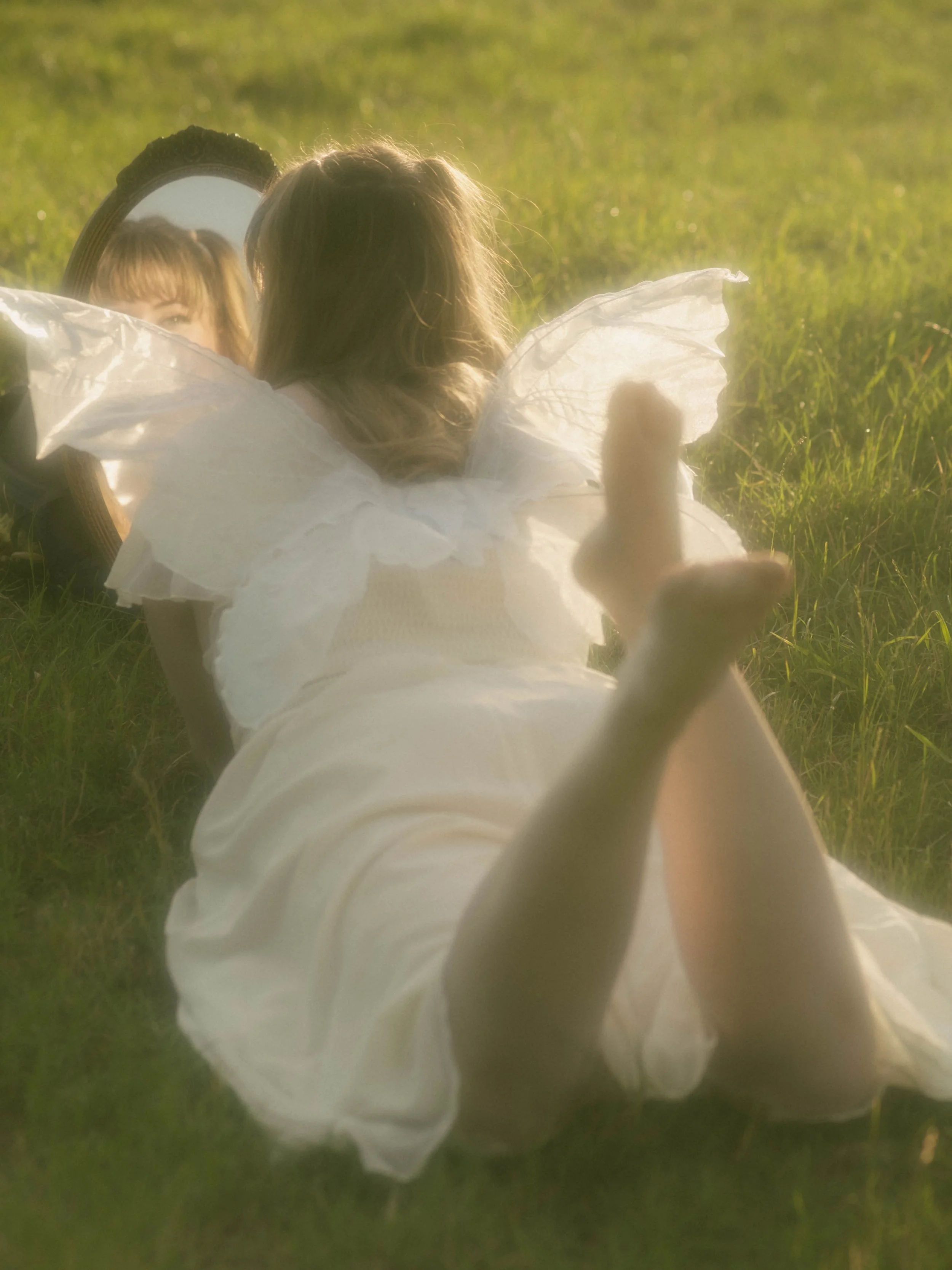 A woman dressed as a fairy with transparent wings lying on grass, looking into a mirror on the ground that reflects her face.