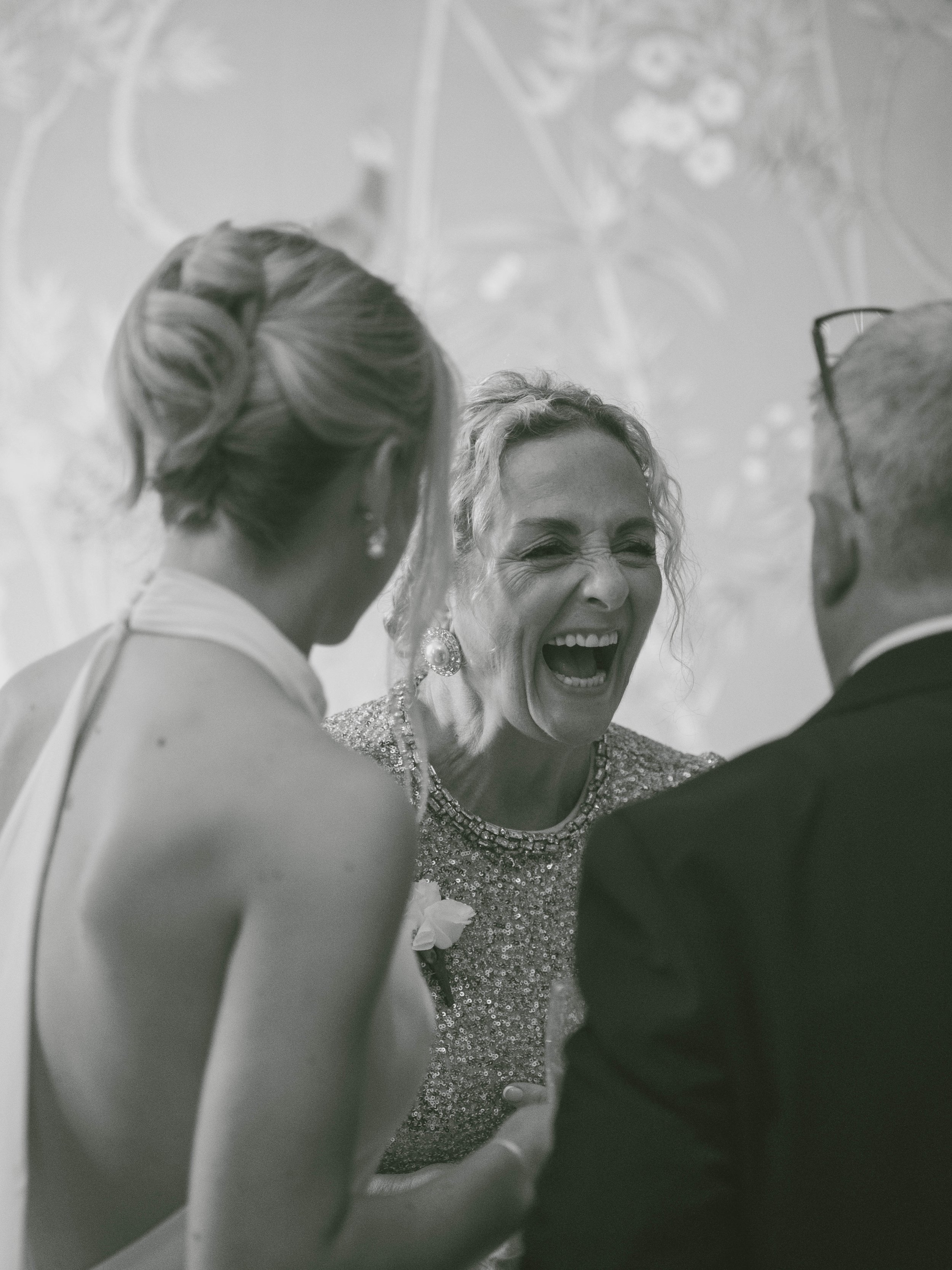 Three women, one with curly hair and earrings, in formal attire, smiling and looking at each other, with a decorative backdrop.