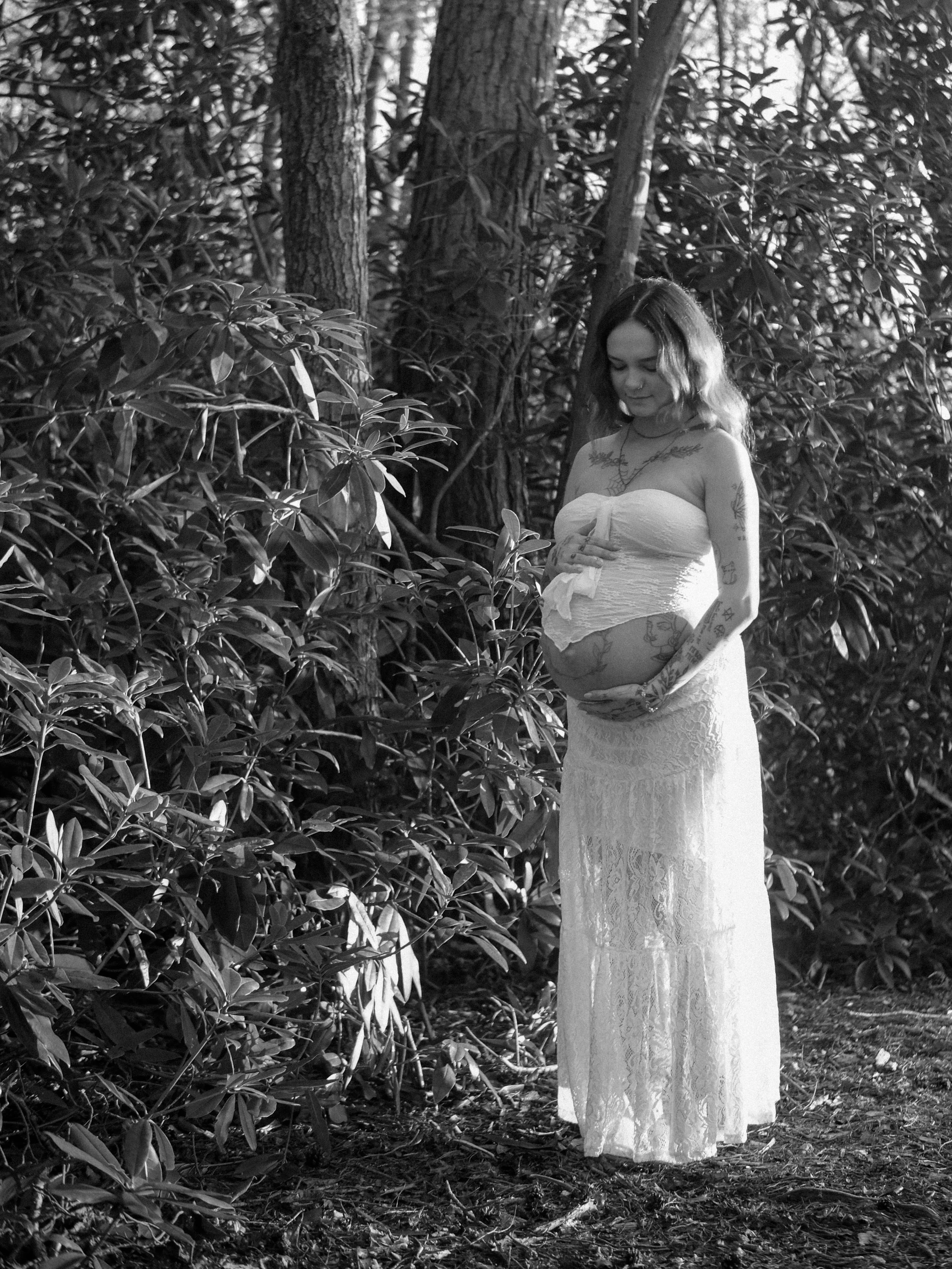A woman in a white dress with tattoos is standing among trees and bushes in a forest, looking down and holding her stomach. The image is in black and white.