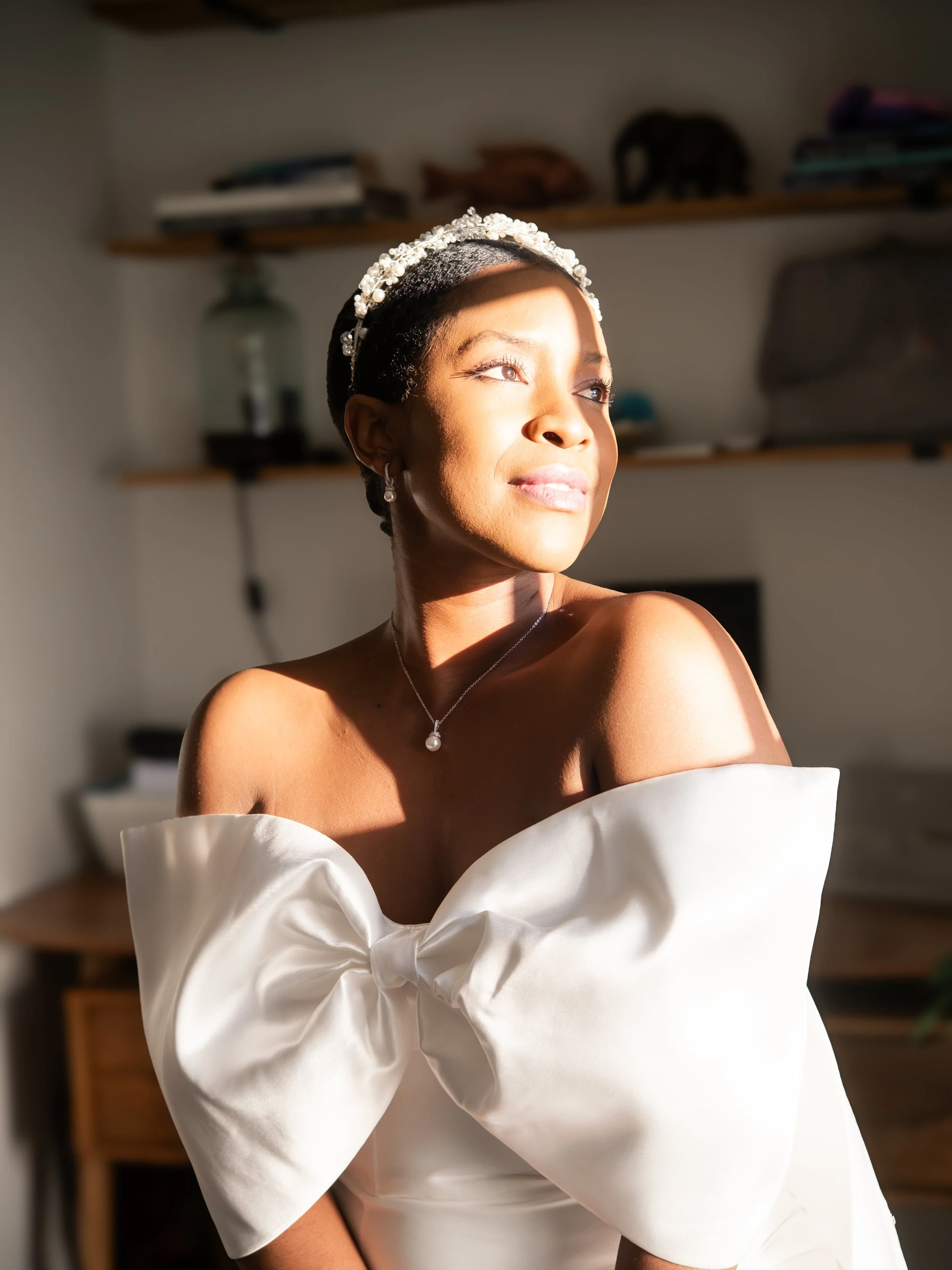 A woman wearing a pearl headband and pearl earrings, dressed in an elegant off-the-shoulder white gown, standing in a sunlit room.