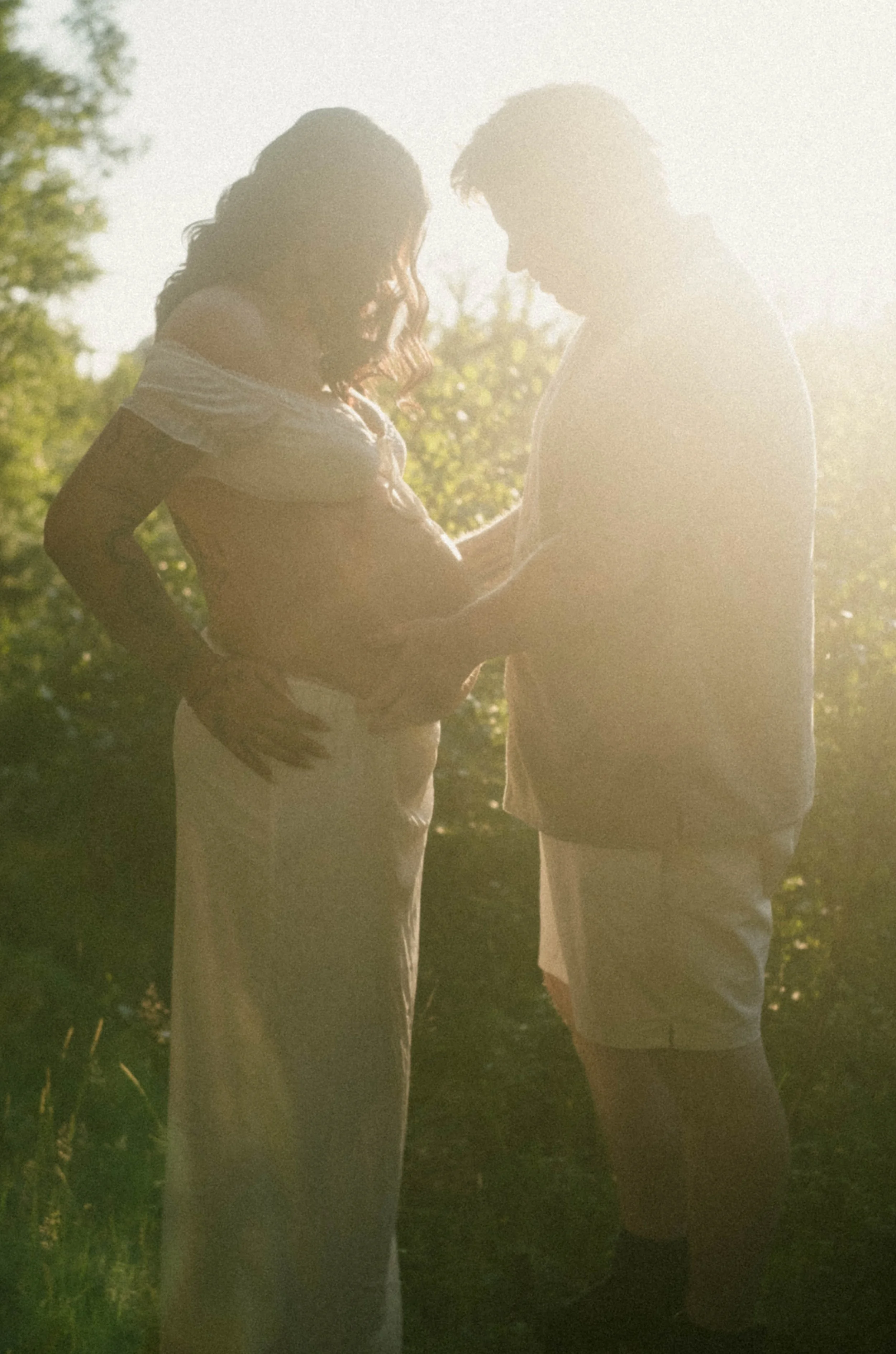 A couple standing close together outdoors during sunset or sunrise, with the woman holding the man's hands and both looking at each other, surrounded by trees and greenery.