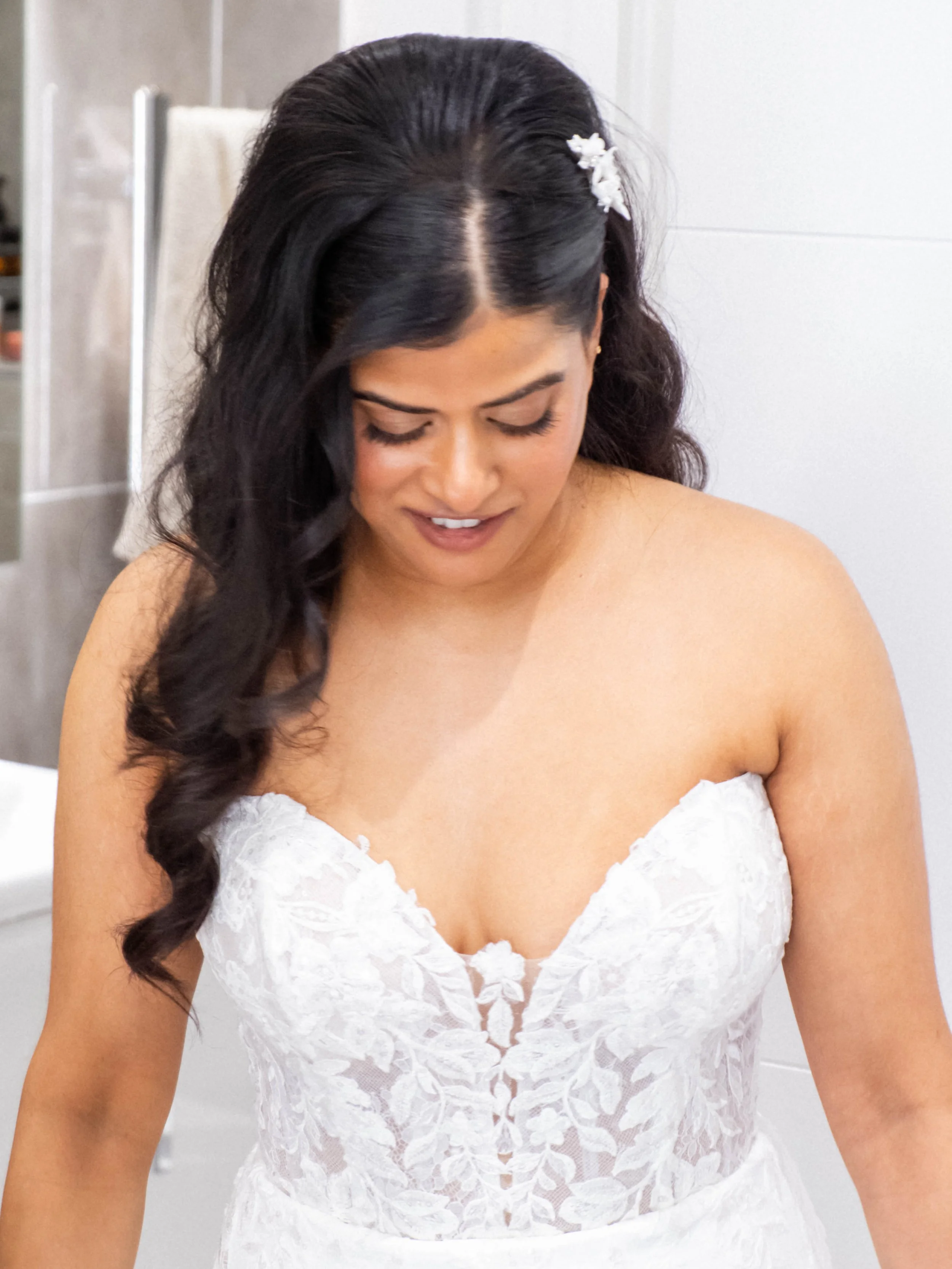 A woman with dark, wavy hair styled with a white floral hair accessory, wearing a white lace strapless dress, smiling downwards. The background includes a white wall and a towel rack.