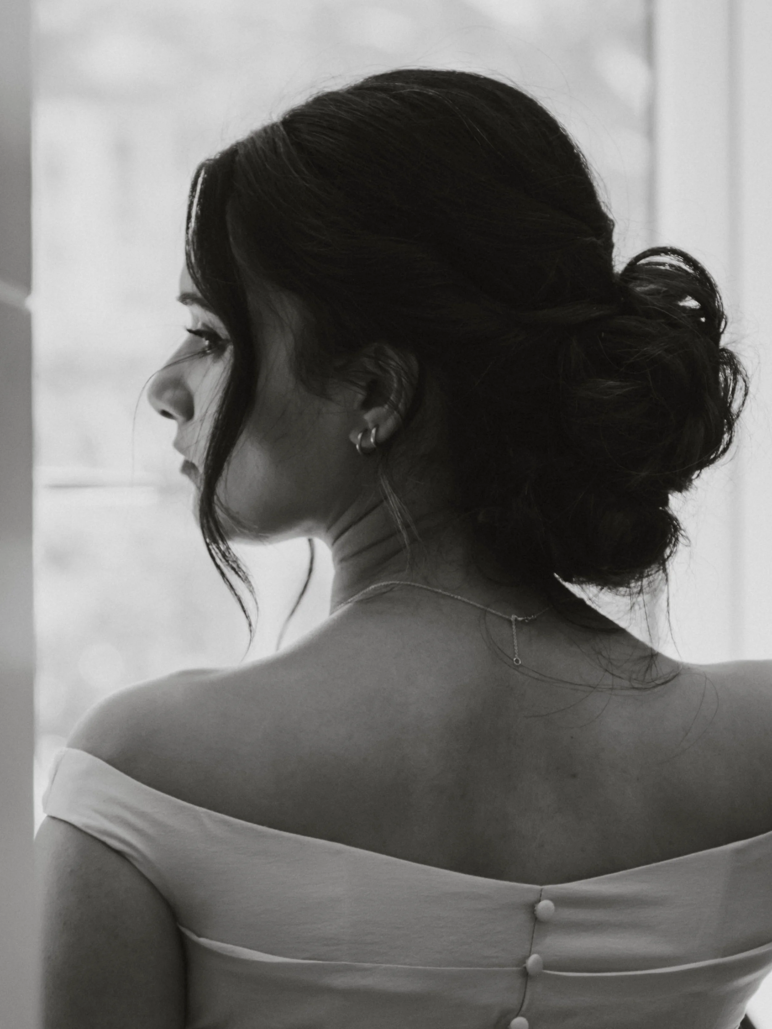 Black and white photo of a woman with dark hair styled in an updo, looking to the side, wearing a dress with button details on the back.