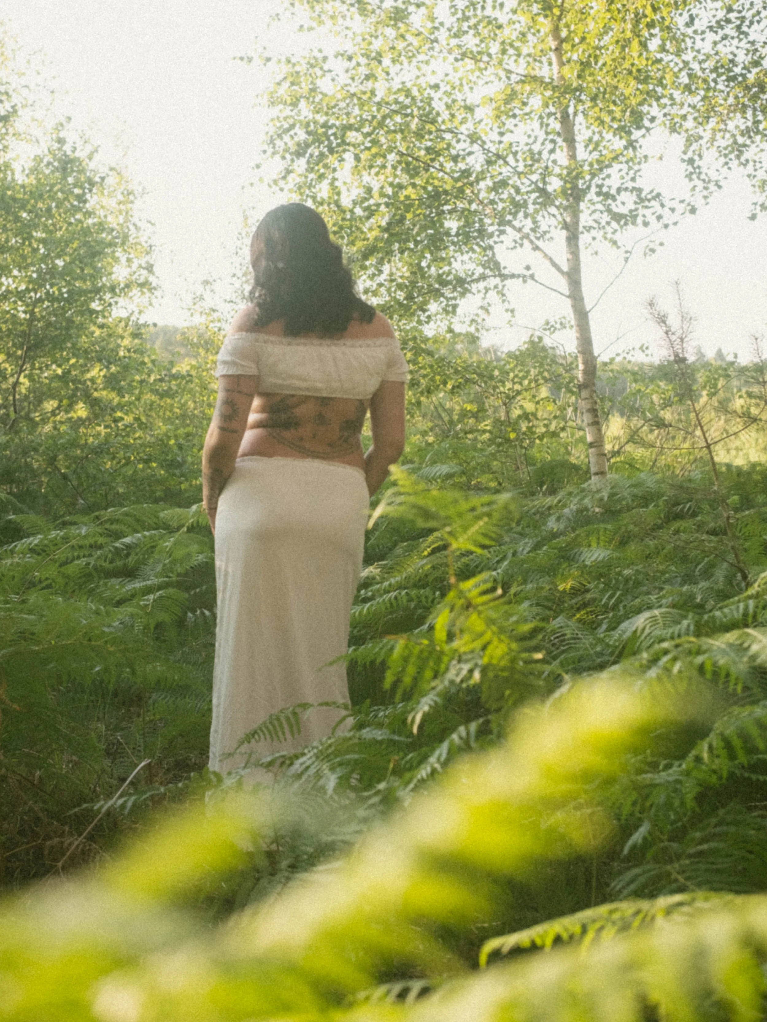 A woman with tattoos on her arms and back, wearing a white off-shoulder top and a long white skirt, standing in a lush green forest with sunlight filtering through the trees.