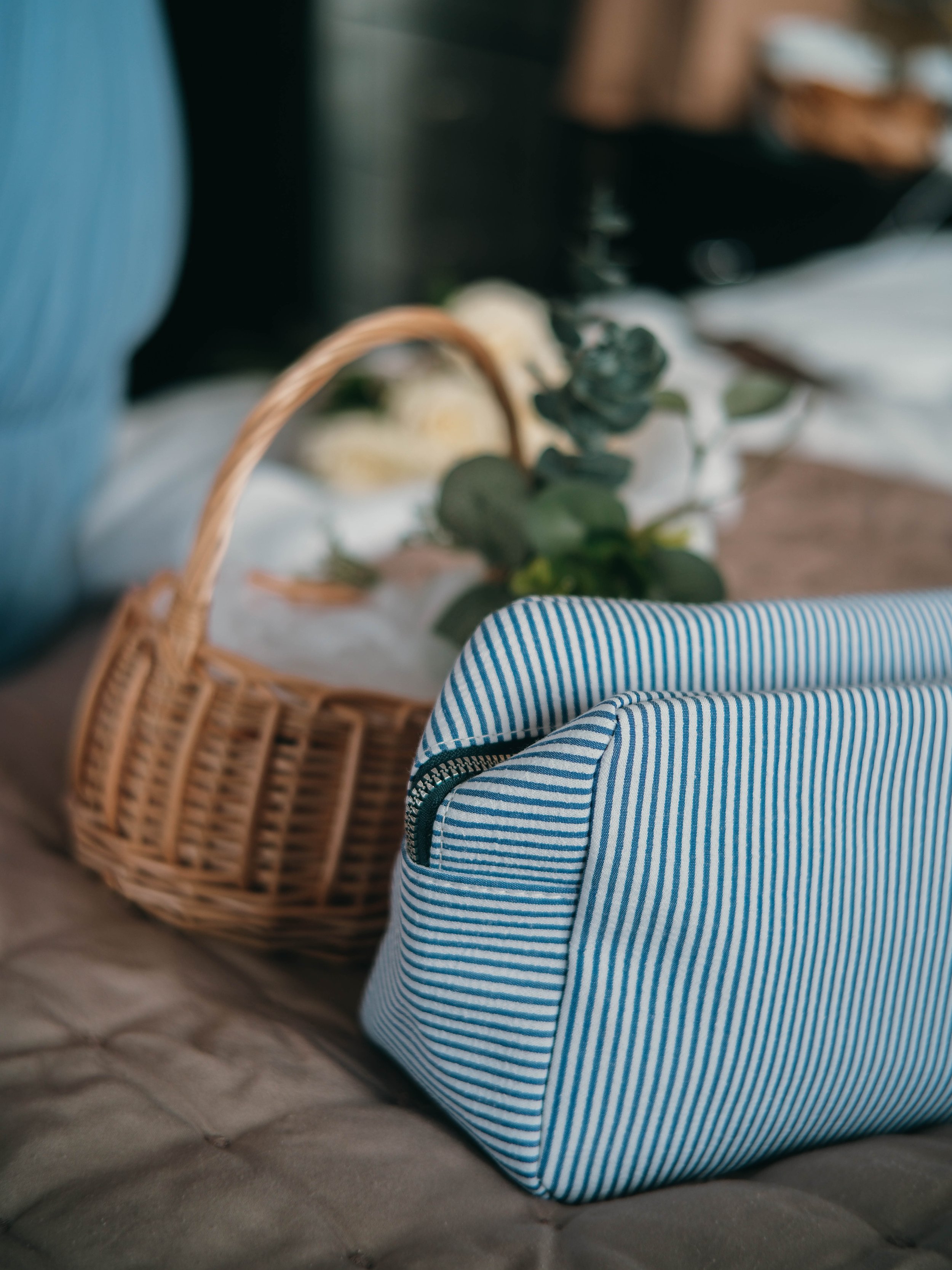 Close-up of a blue and white striped fabric bag on a quilted surface, with a wicker basket filled with flowers and greenery nearby.