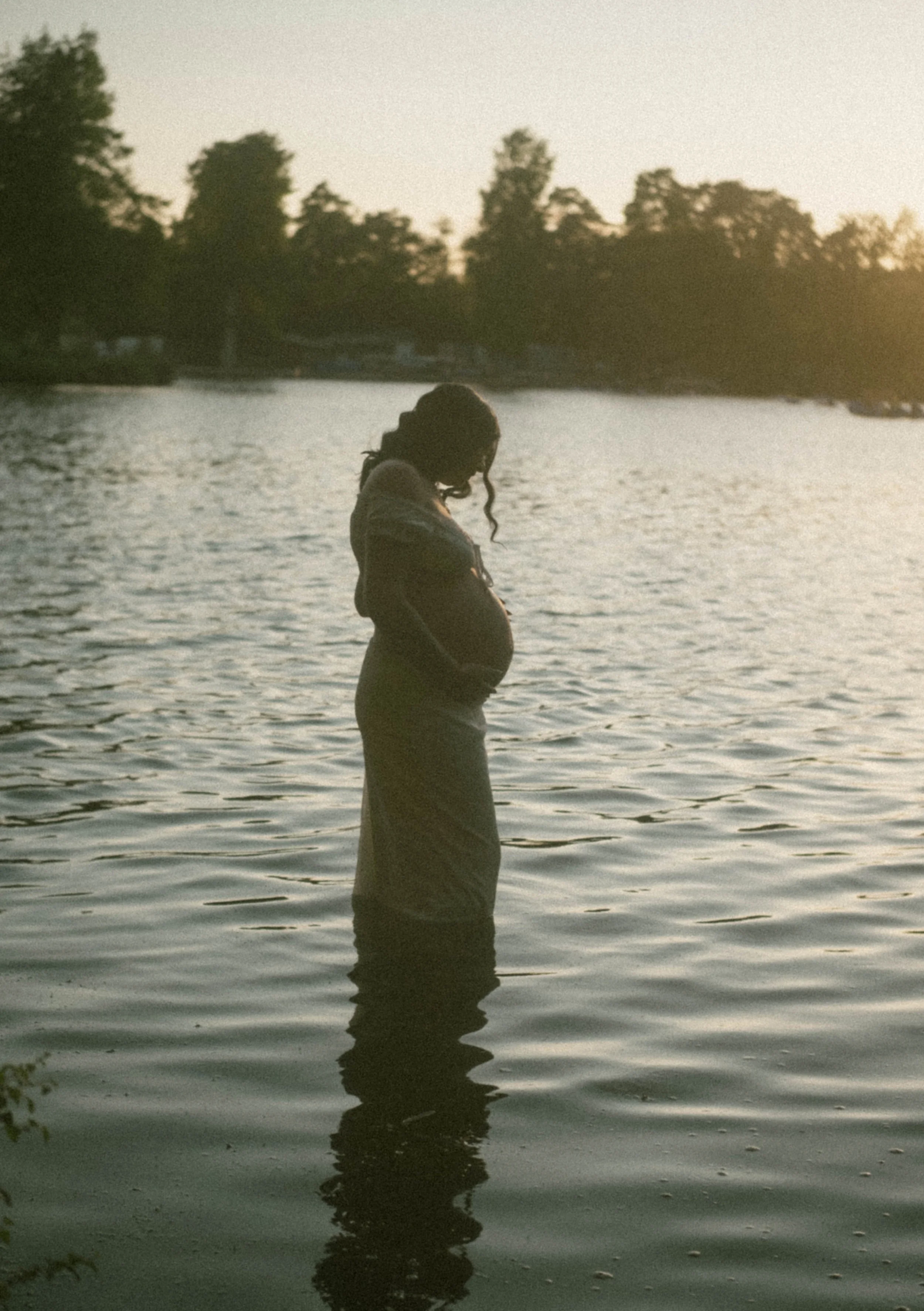 A woman standing in a body of water at sunset, with trees in the background and her reflection visible in the water.