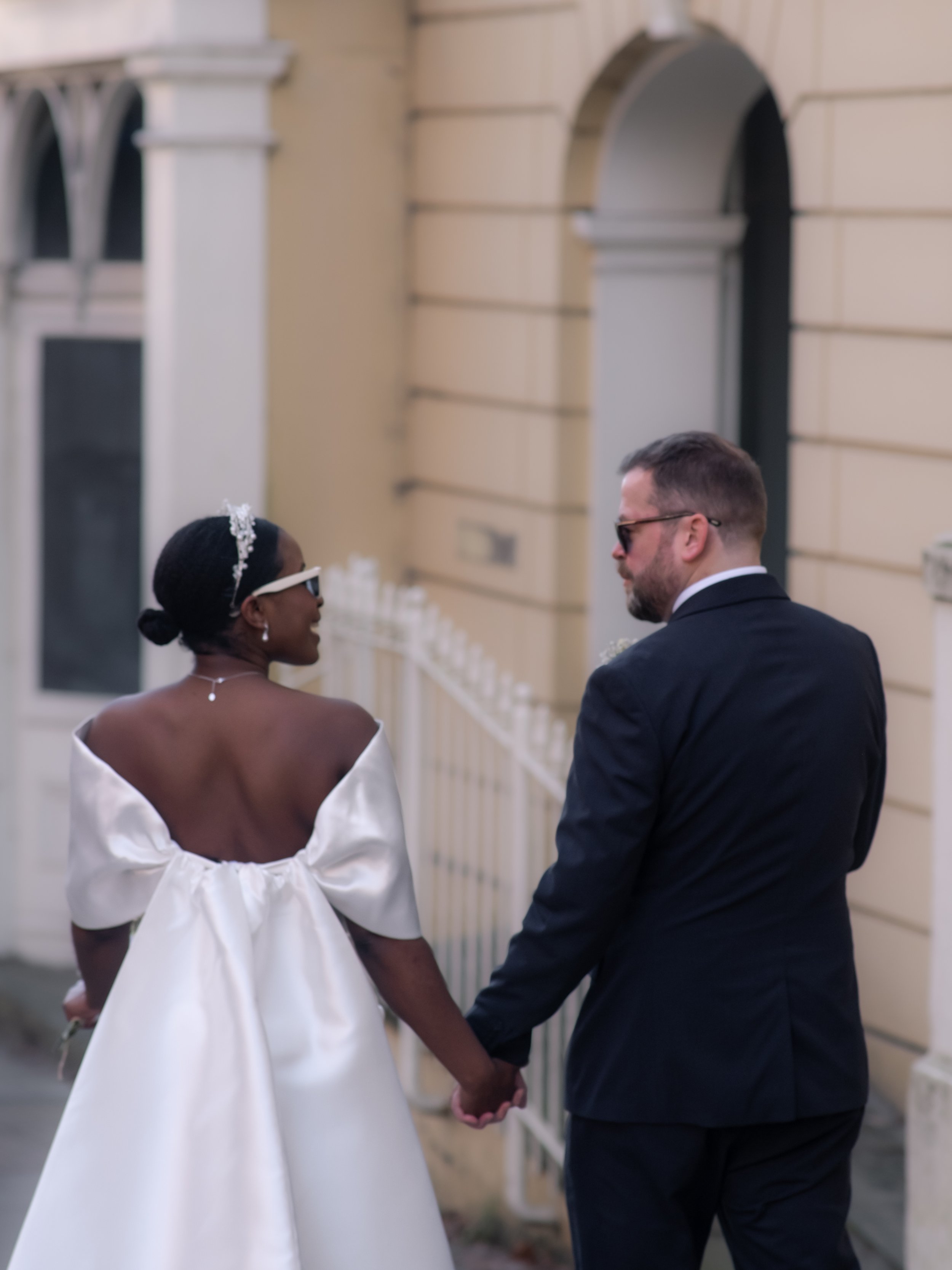 A couple holding hands and facing each other, with the woman wearing a white off-shoulder dress and a tiara in her hair, and the man wearing a dark suit and sunglasses, standing outside a building with architectural details.