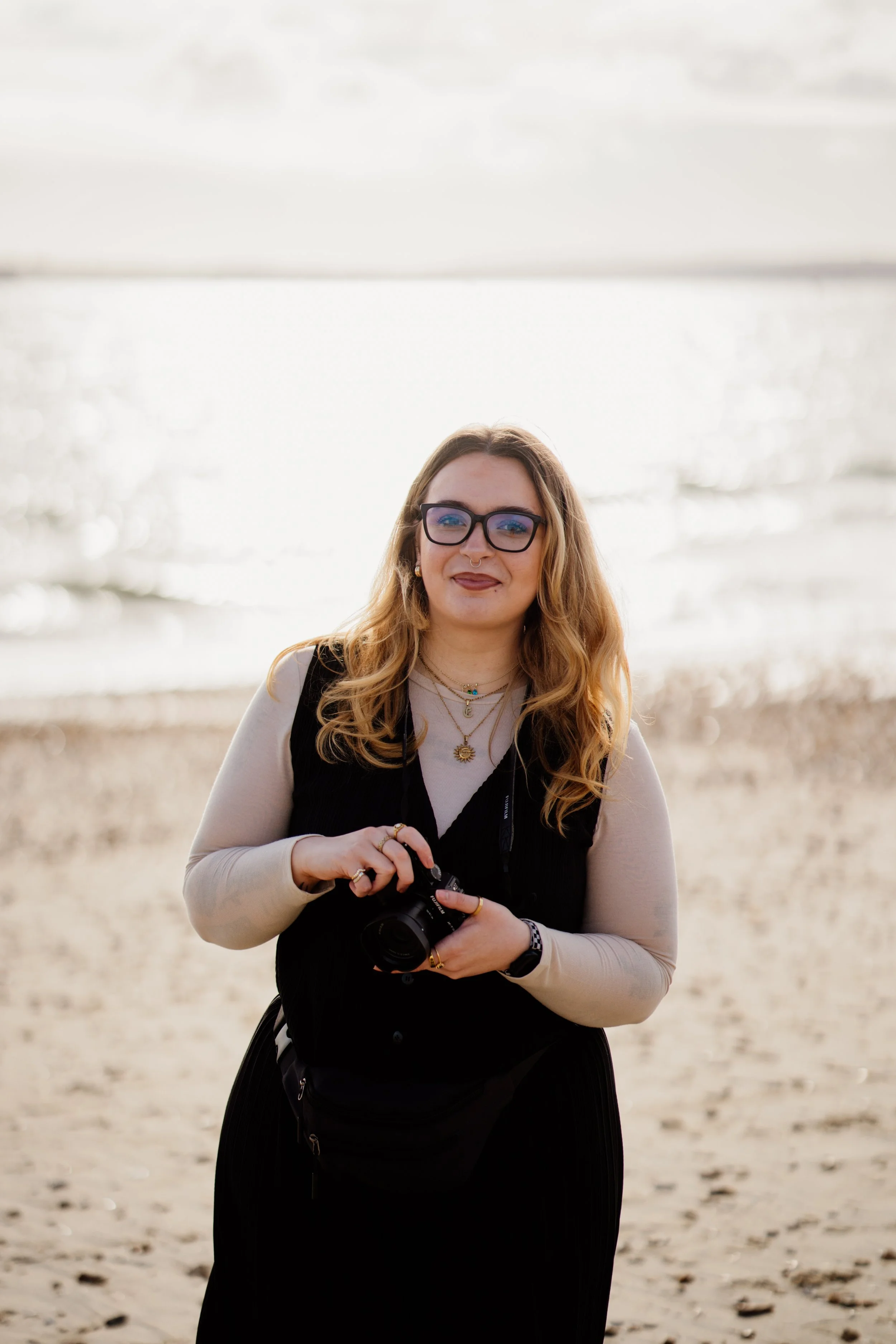 A woman with long wavy hair wearing glasses, a beige long-sleeve shirt, and layered necklaces, holding a camera, standing on a sandy beach with the ocean and cloudy sky in the background.