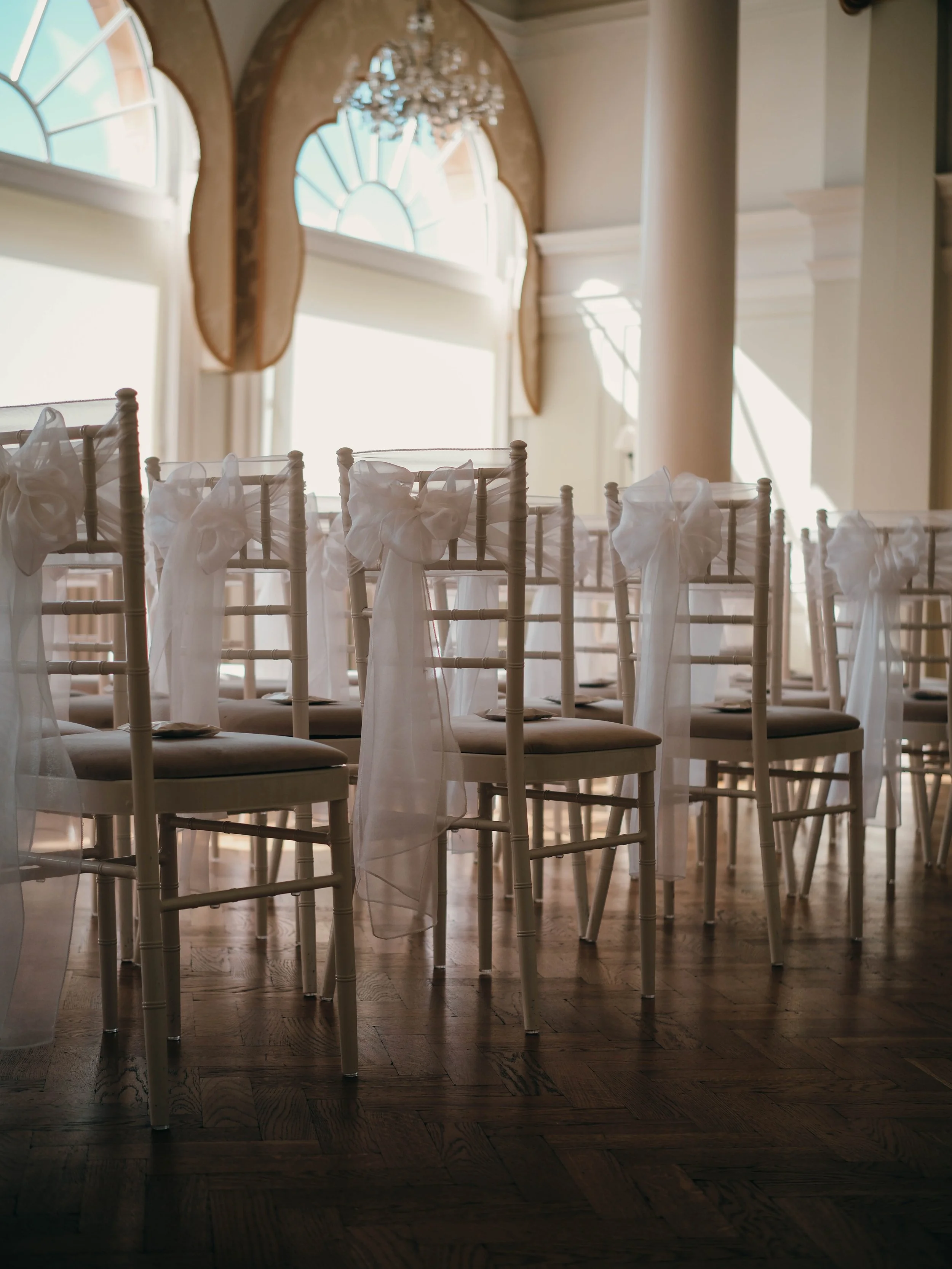 Elegant event space with white chairs tied with white sashes, set up for a wedding or similar celebration, with large windows and natural light highlighting the wooden floor and drapery.