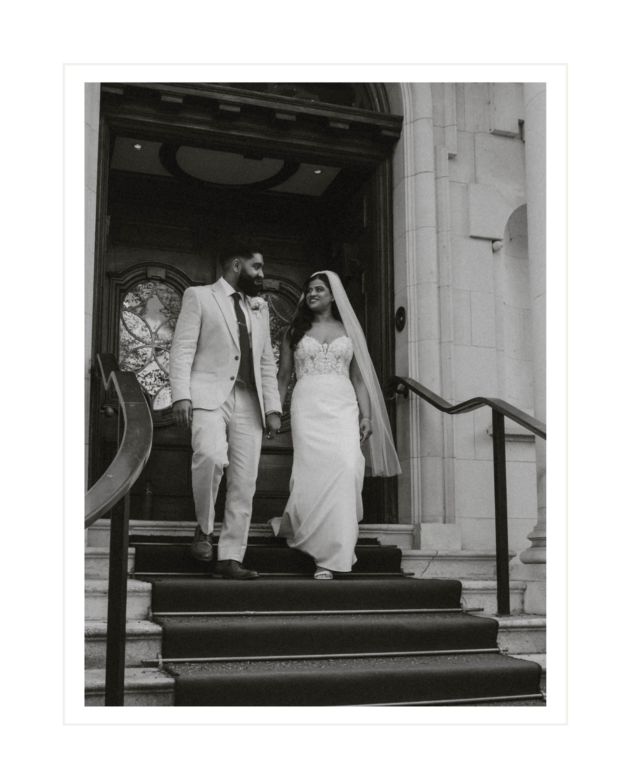A black and white photograph of a newlywed couple standing on the steps of a building with ornate woodwork and glass doors. The groom, wearing a light suit and tie, is looking at the bride with a smile. The bride, in a lace wedding dress with a veil, is looking at the groom with a smile. They are holding hands, and the bride's dress trails slightly on the steps.