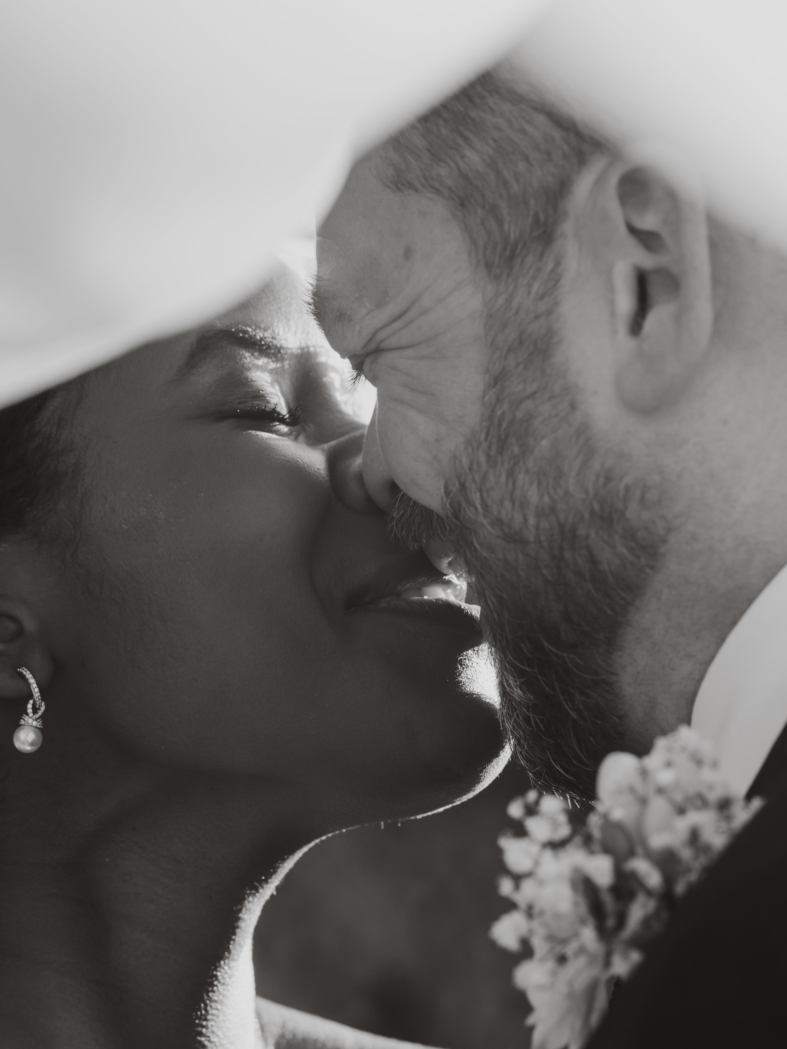 Black and white close-up photo of a couple kissing, with the woman wearing an earring and the man having a beard, against a blurred background of flowers.