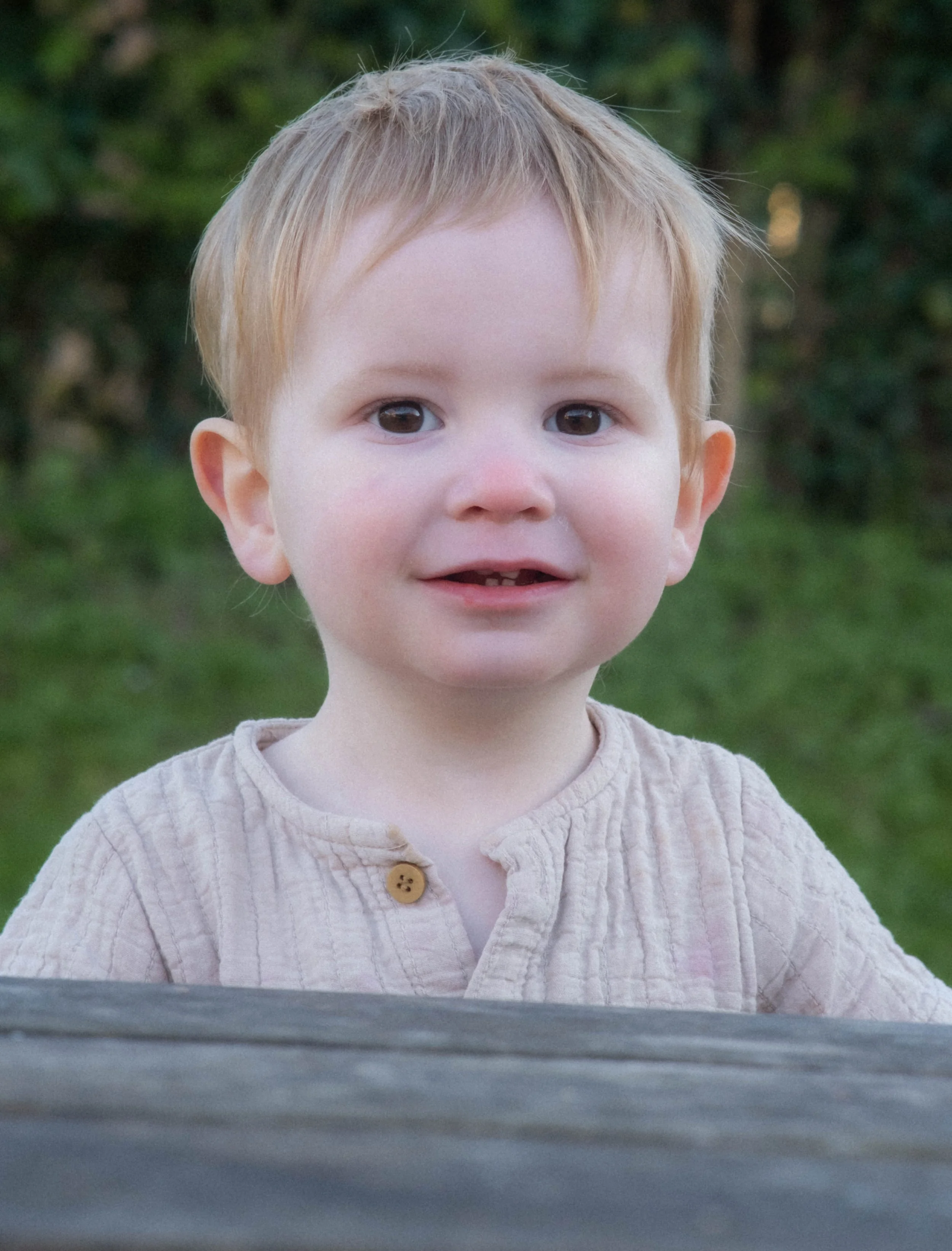 Close-up of a young child with light-colored hair, wearing a beige shirt with a button, outdoors with a green, leafy background.