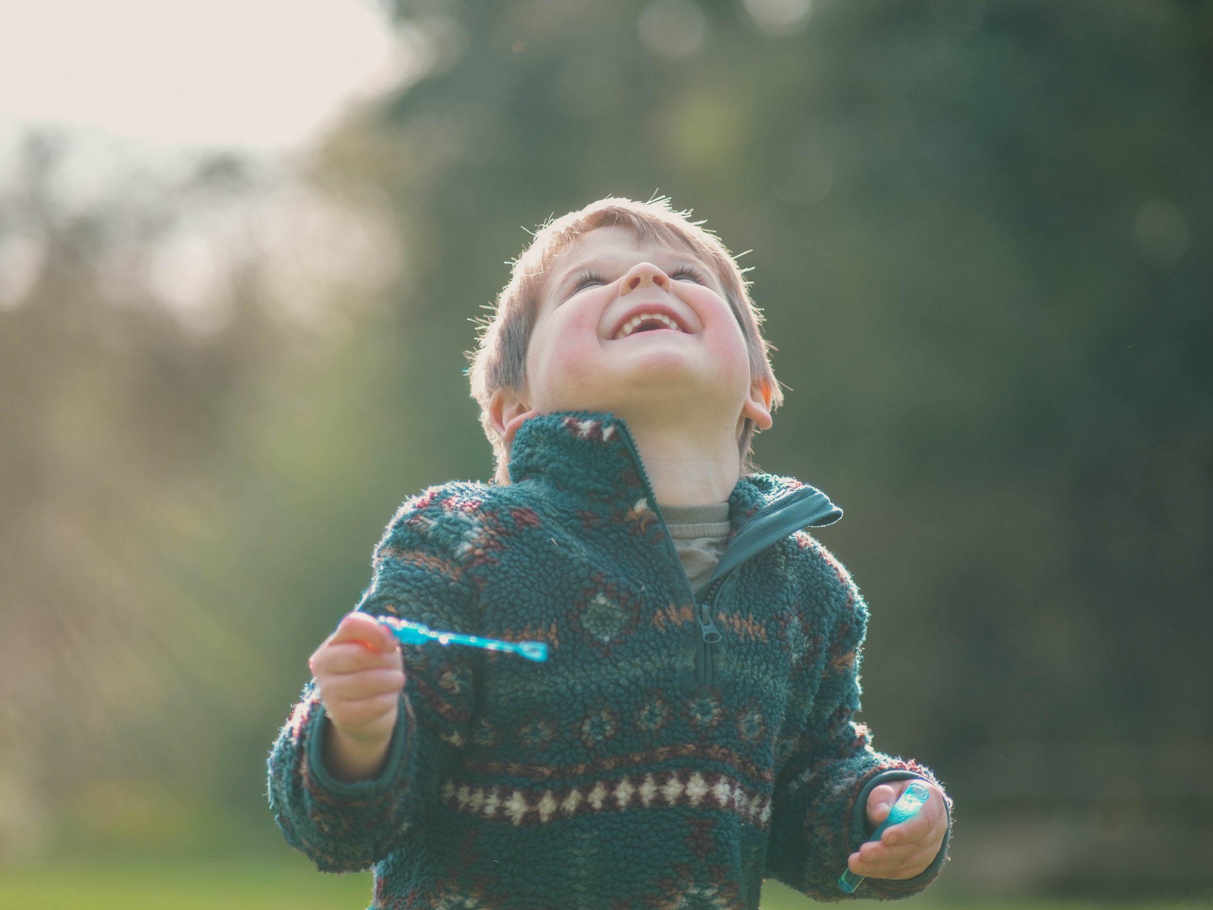 A young boy with short blonde hair is smiling and looking upward while holding a toy sword. He is wearing a patterned fleece jacket and is outdoors with blurred green trees in the background.