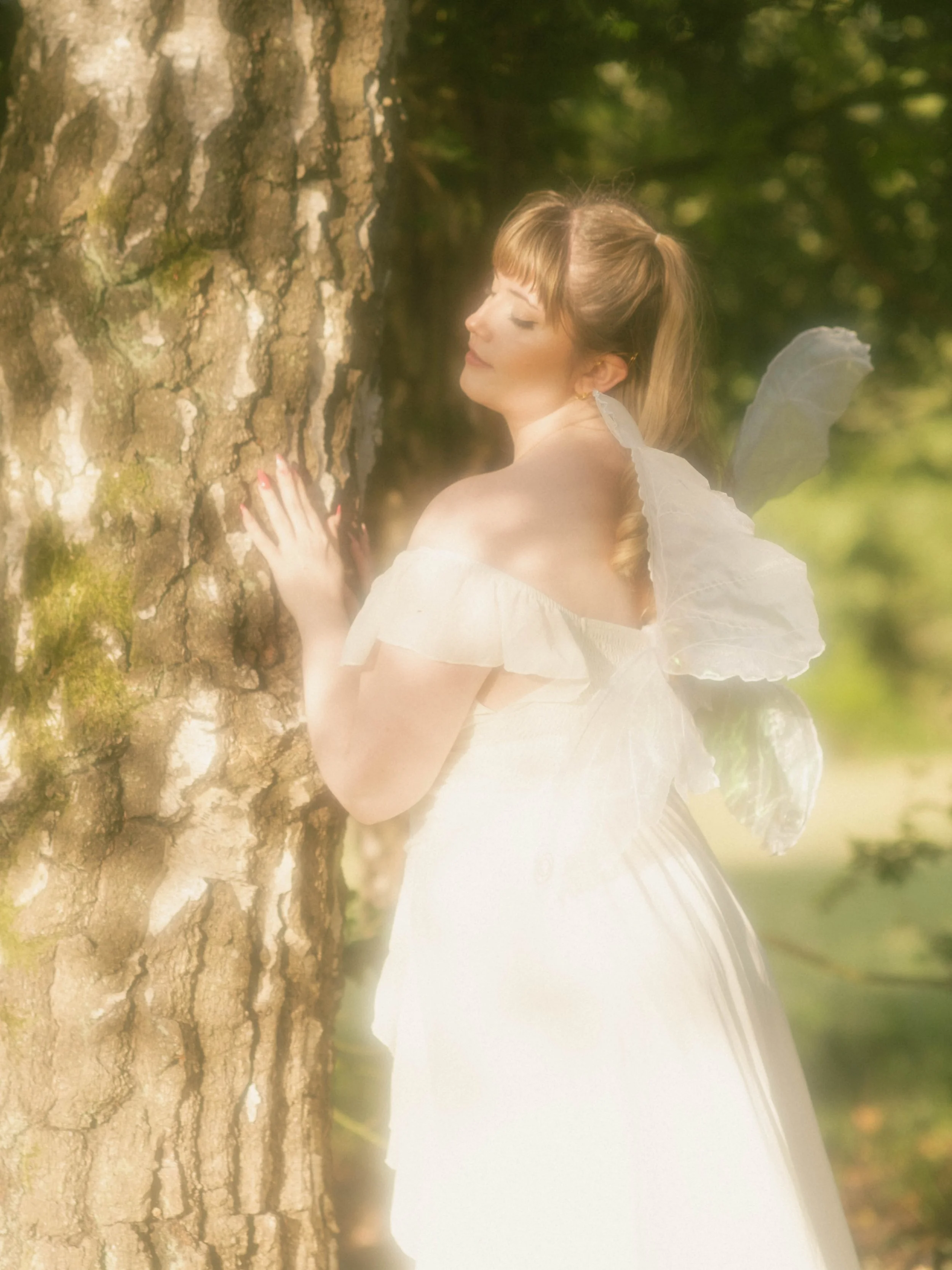A woman dressed as a fairy, with wings and a white dress, hugs a large tree trunk in a forested area on a sunny day.