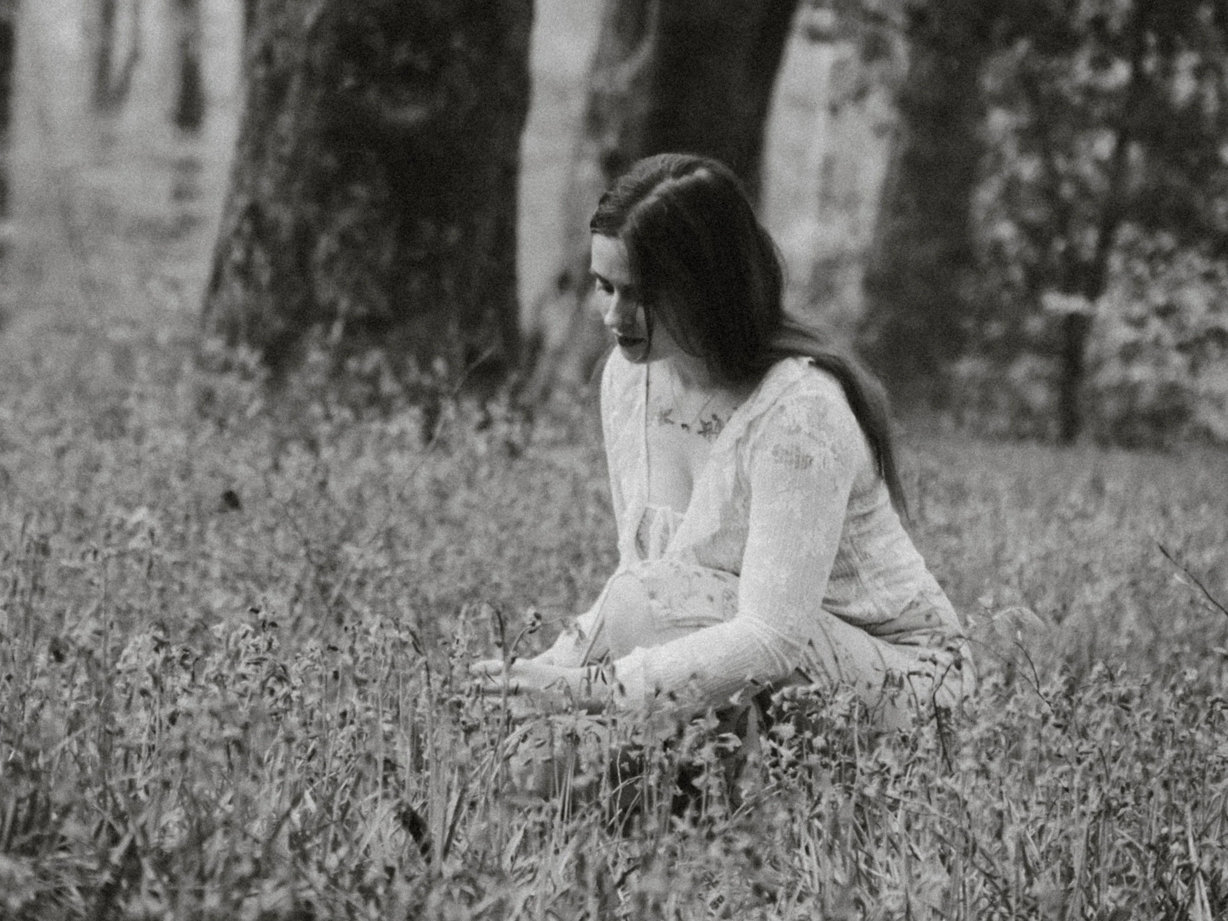 A woman with long dark hair, dressed in traditional Asian attire, sits on the grass outdoors, surrounded by trees and nature.