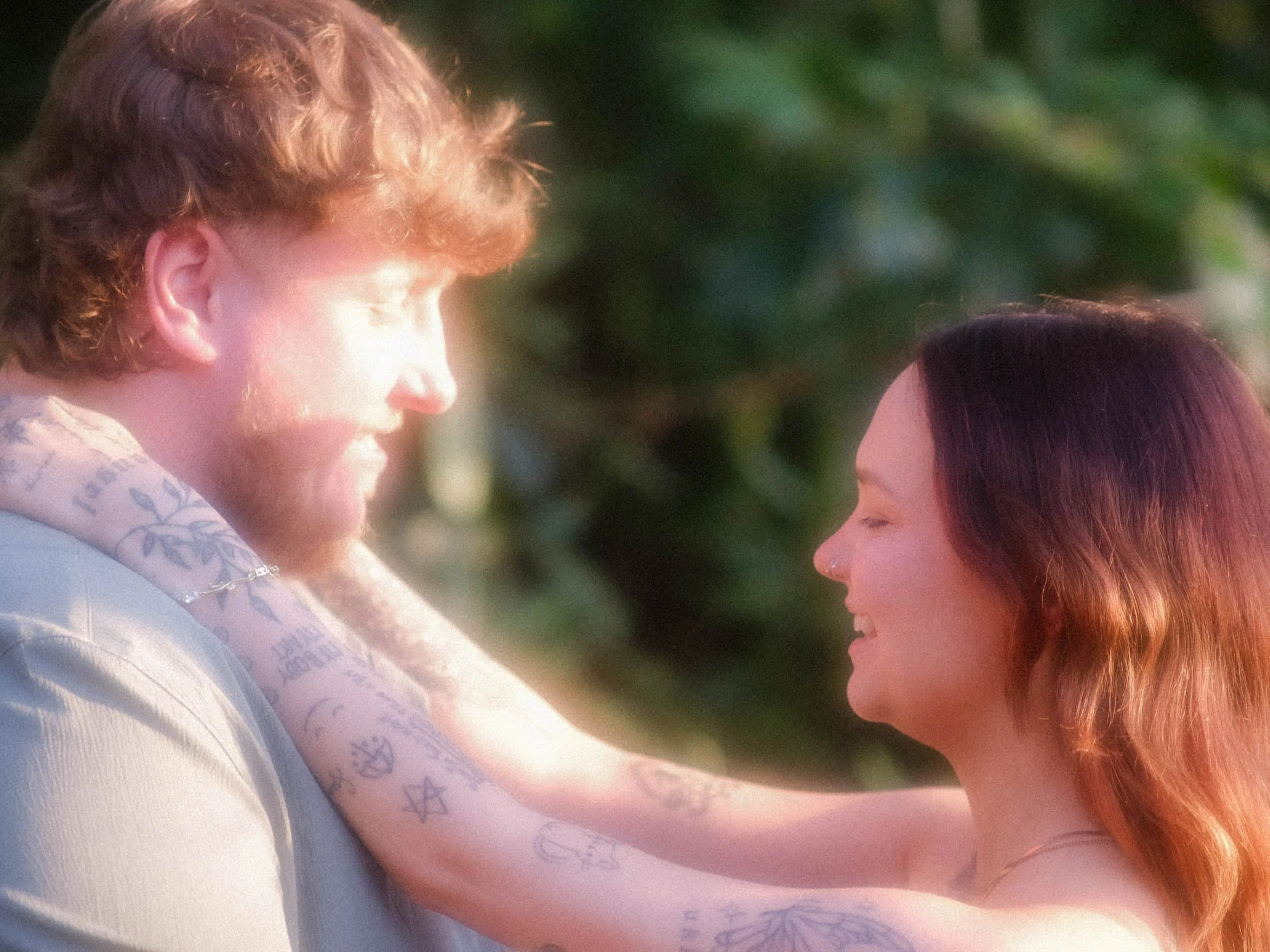 A man with reddish hair looking at a woman with long red hair, both appearing to be in an outdoor setting with green foliage in the background.
