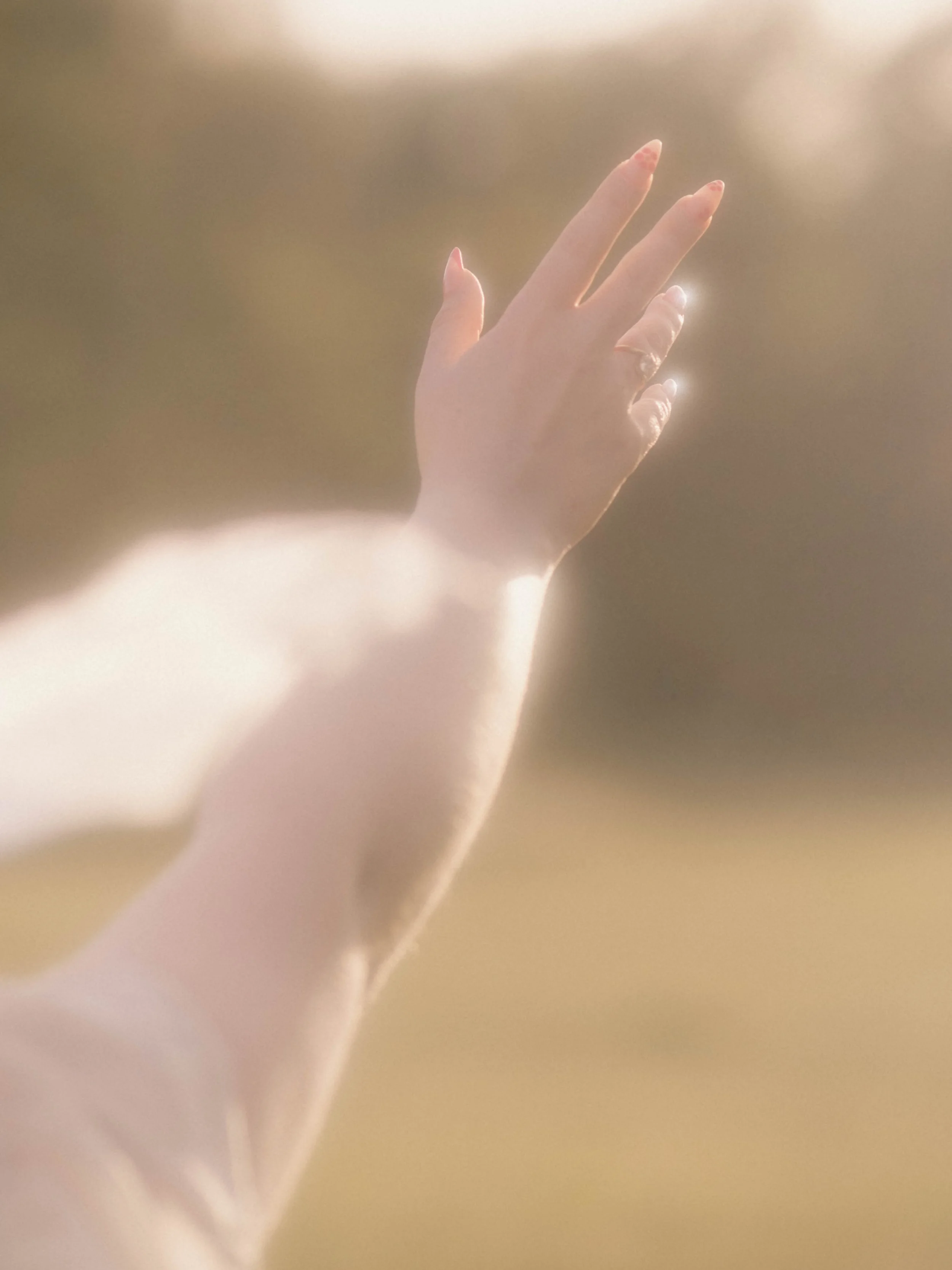 A person's hand reaching upward with fingers slightly spread, against a softly lit, blurred background.