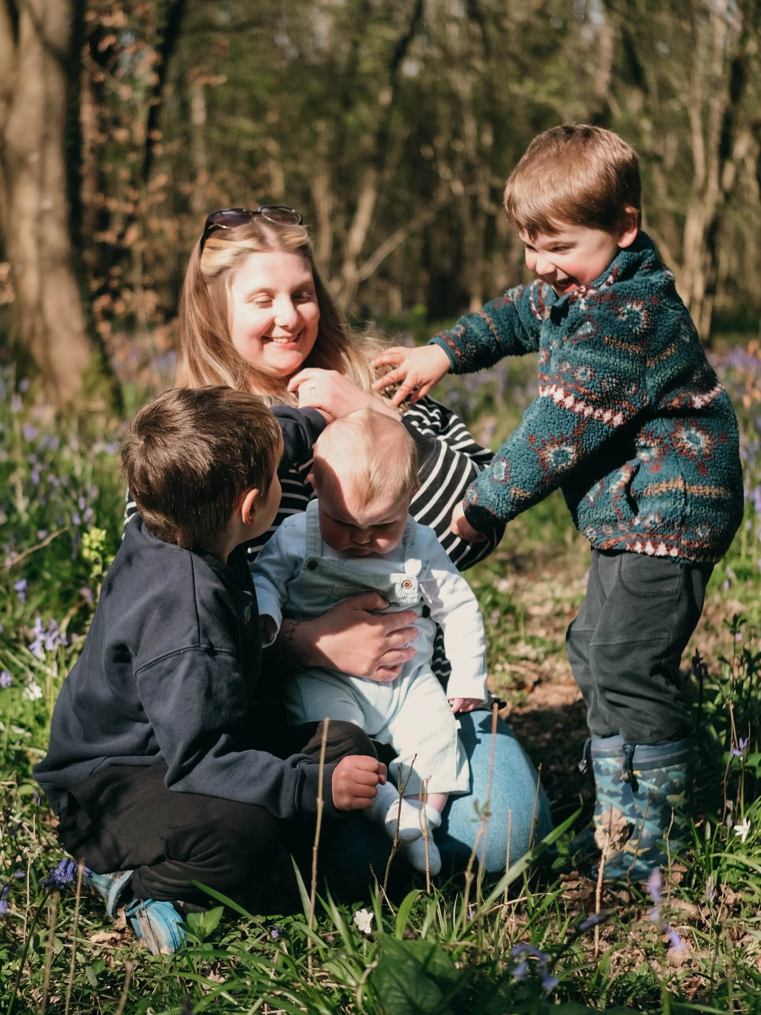 A woman smiling and playing with children outdoors in a wooded area during spring, surrounded by flowers and trees.