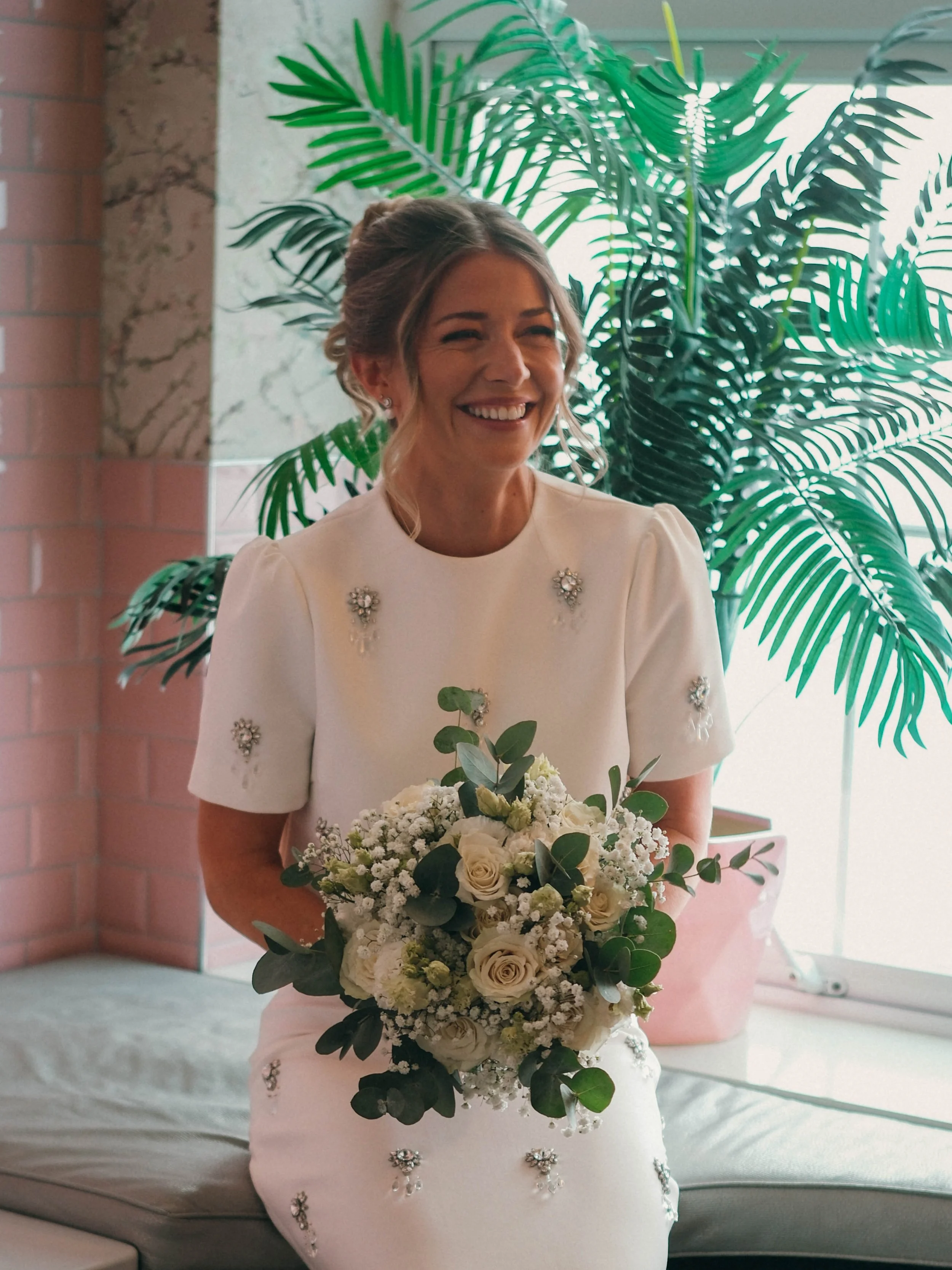 A smiling woman in a white dress holding a bouquet of white and green flowers, standing indoors near a large green leafy plant.