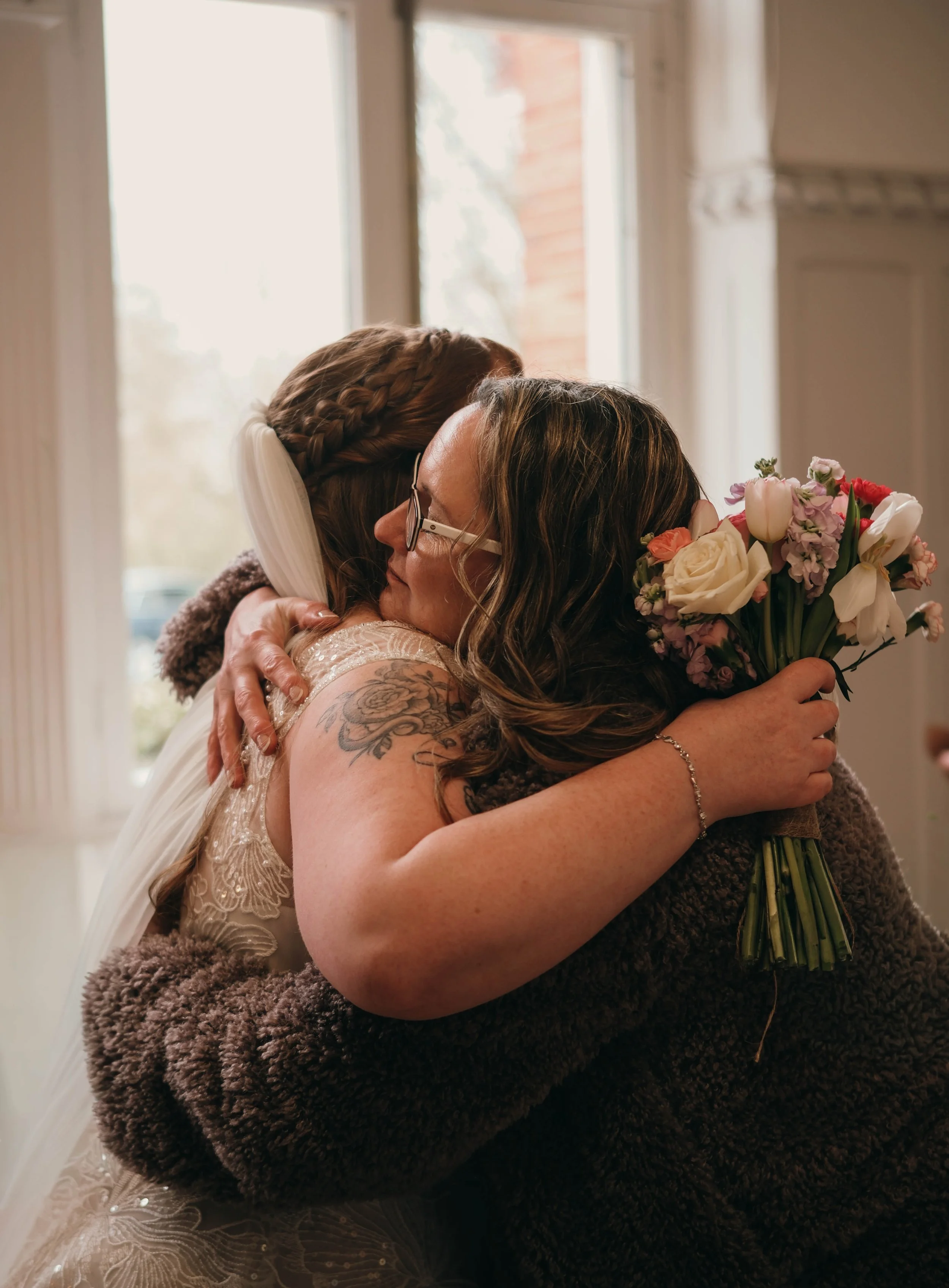 Two women hugging, one holding a bouquet of flowers, inside a room with large windows.