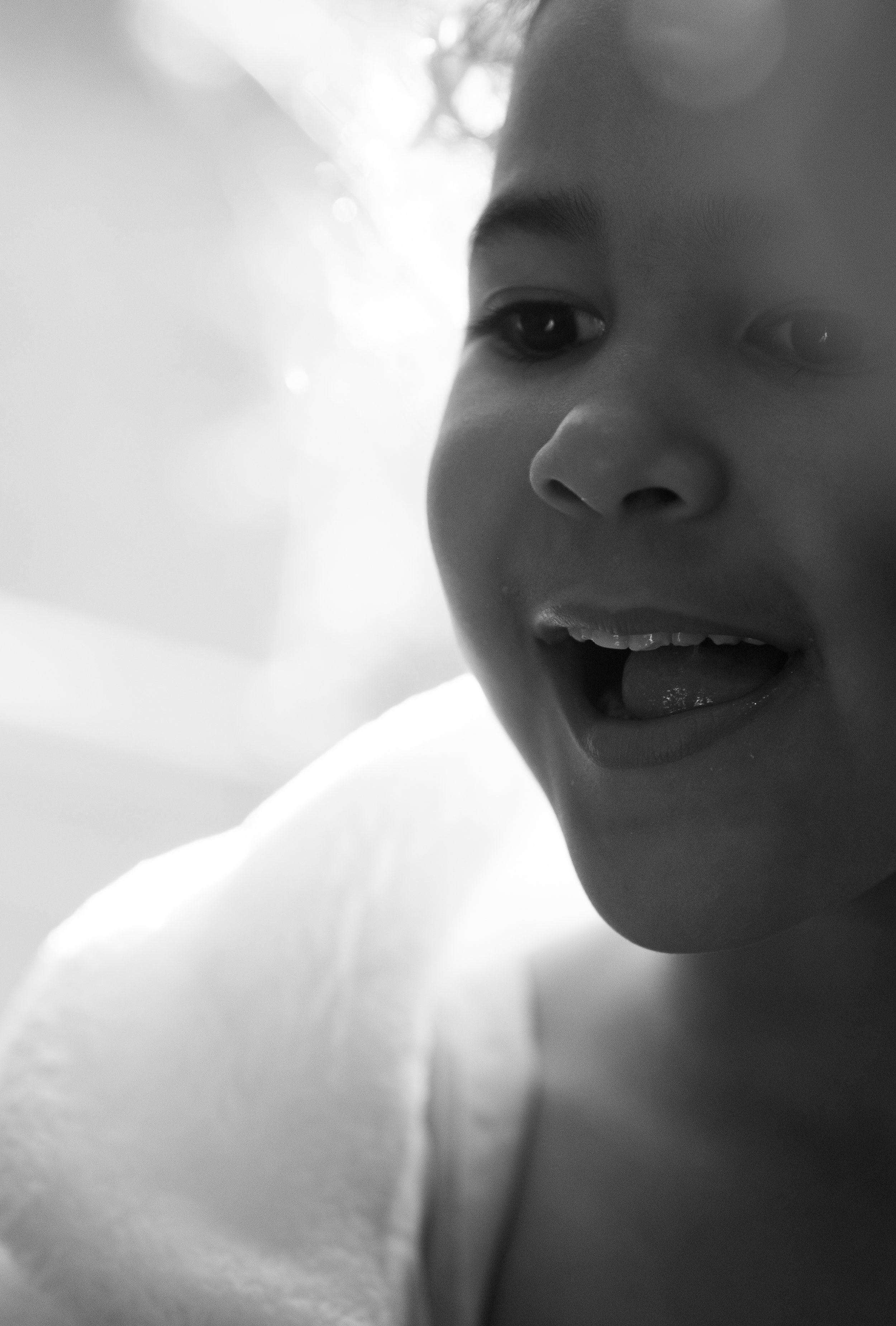 Close-up black-and-white photo of a smiling child's face with their mouth open, showing part of their teeth and tongue, capturing a joyful expression.