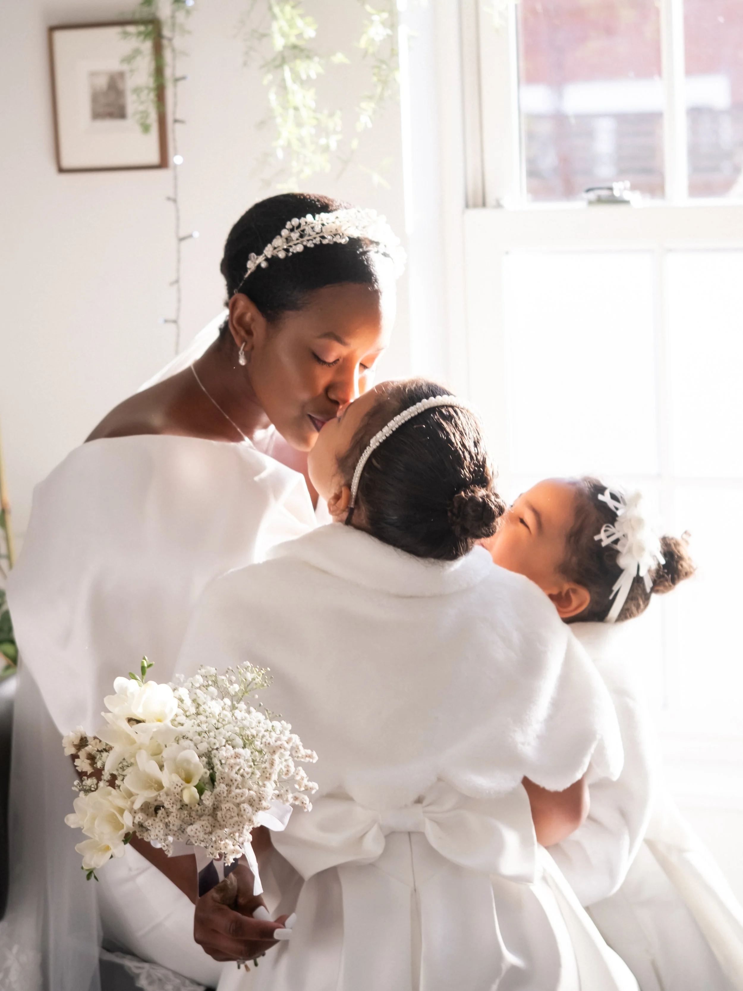 A bride and a young girl share a tender moment, with the bride gently touching the girl's head as they exchange a kiss or whisper. The bride is dressed in a white gown, wearing a bejeweled headband, and holding a bouquet of white flowers. The young g