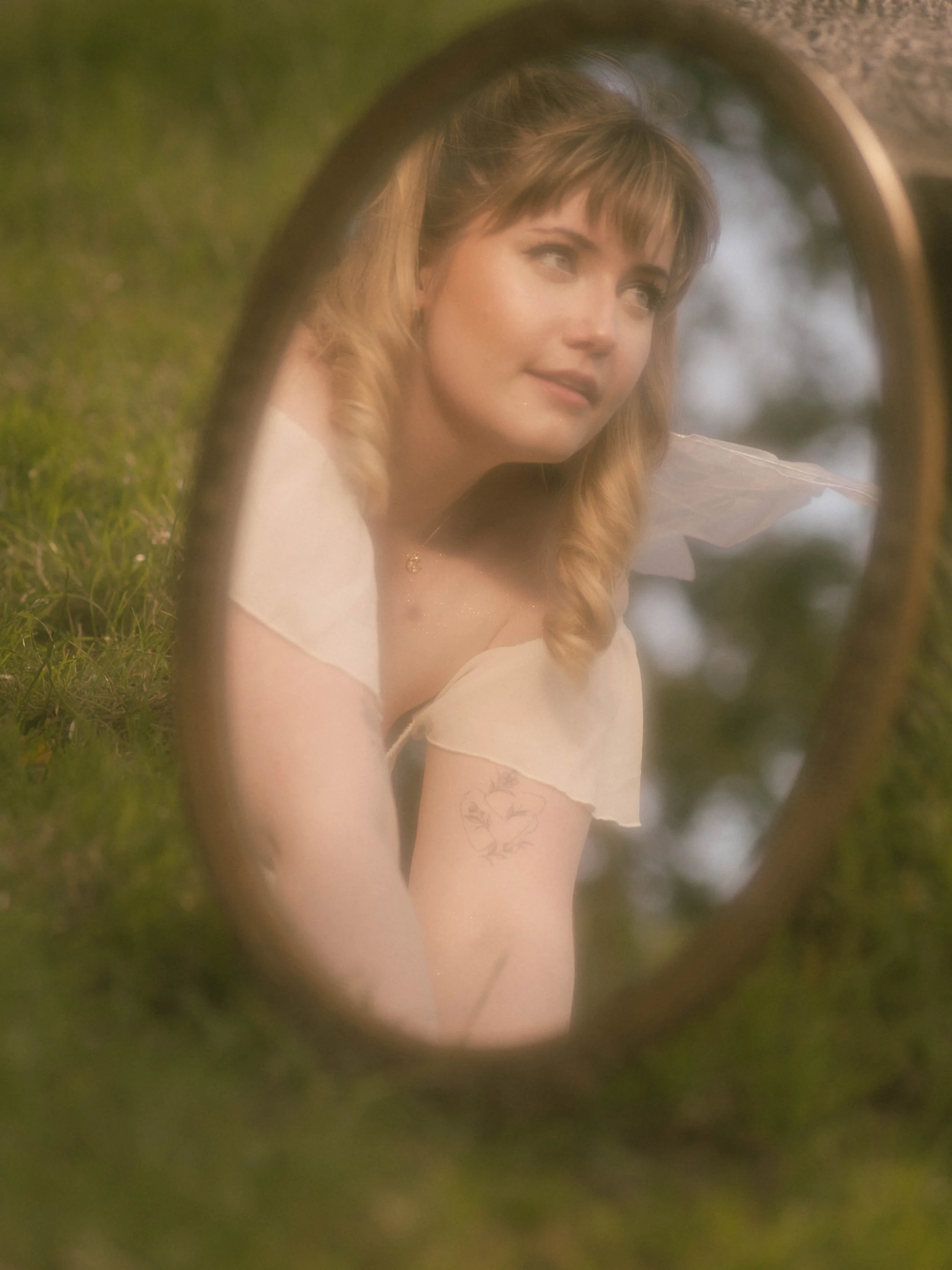 Woman with blonde hair and tattoos looking into a mirror outdoors, framed by the mirror's oval shape, with a blurred background of green foliage and sky.